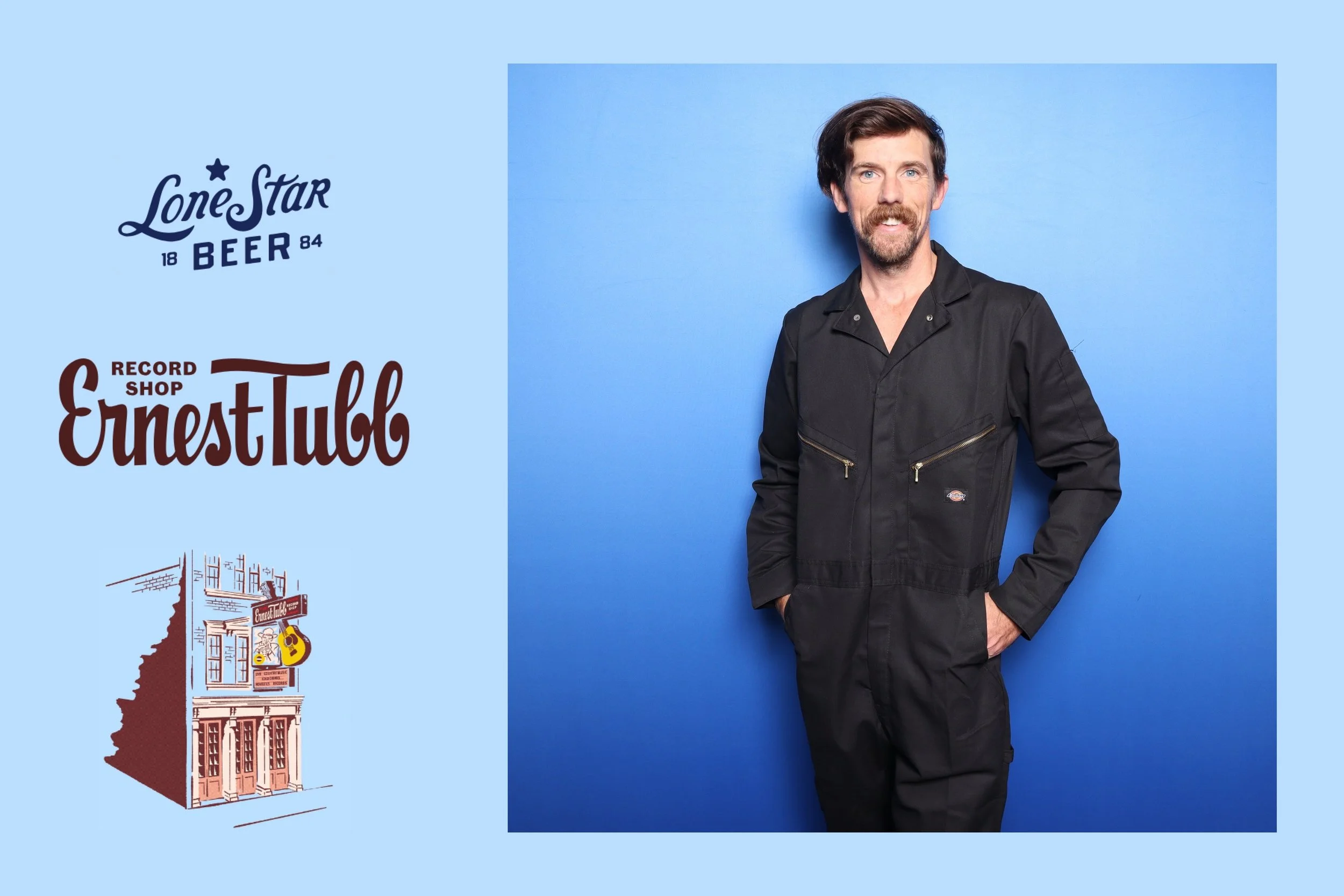 A man with dark hair, beard, and mustache wearing a black work jumpsuit, standing with hands in pockets against a solid blue background.