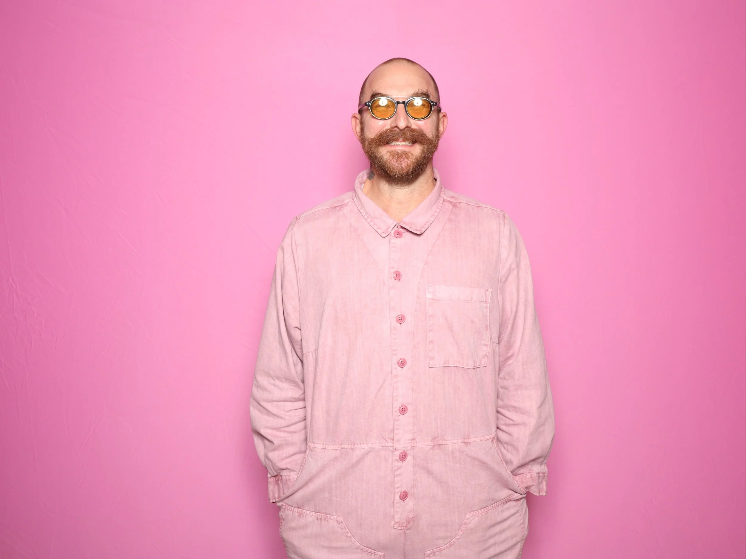 A man with a beard, wearing yellow-tinted sunglasses and a pink shirt, standing against a pink background.