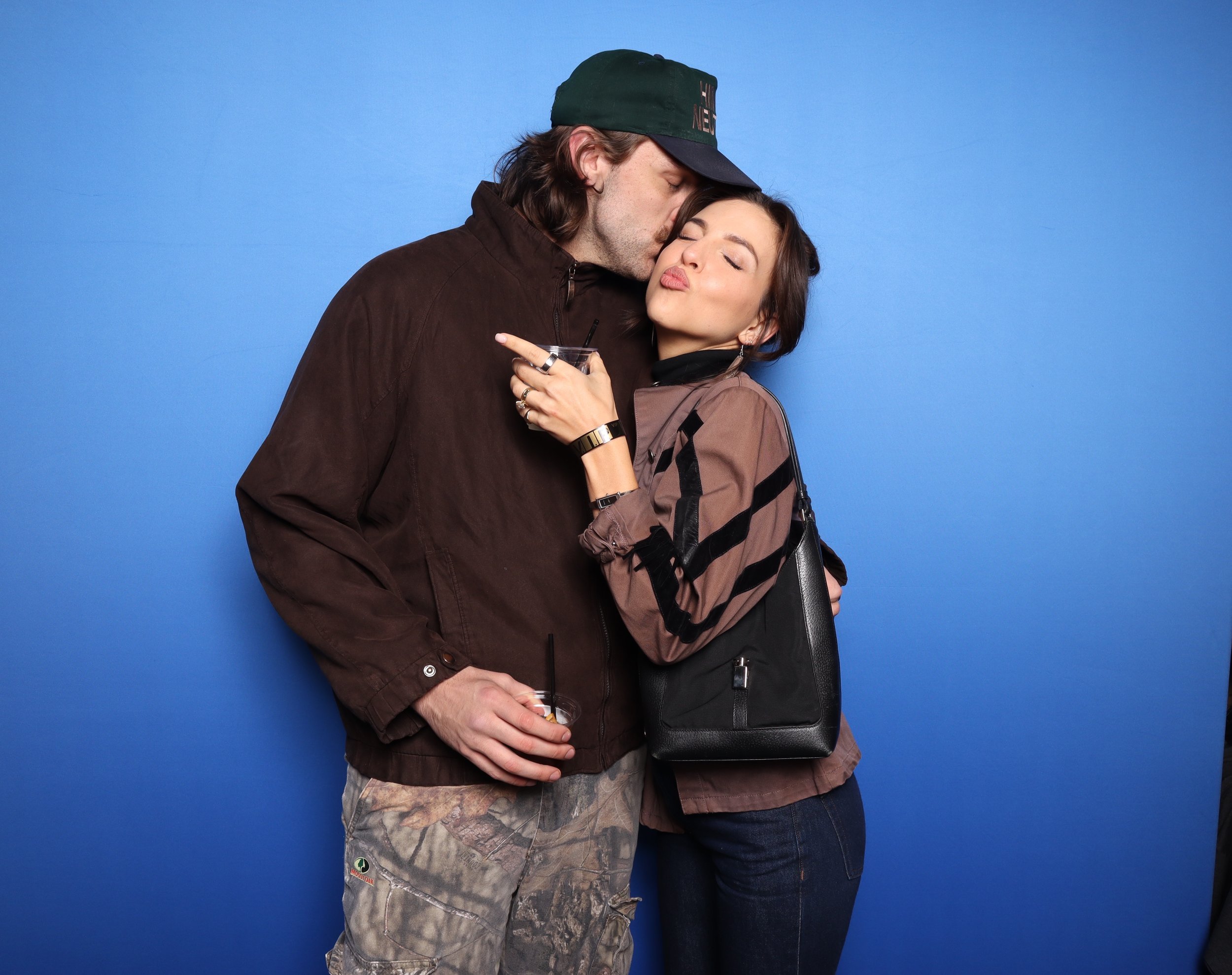 A man and woman hugging against a blue background, with the man giving a kiss on the woman's head. The woman has dark hair, is wearing a brown jacket and a black backpack, and is holding a drink. The man has long hair, is wearing a green cap, a brown jacket, and camo pants.