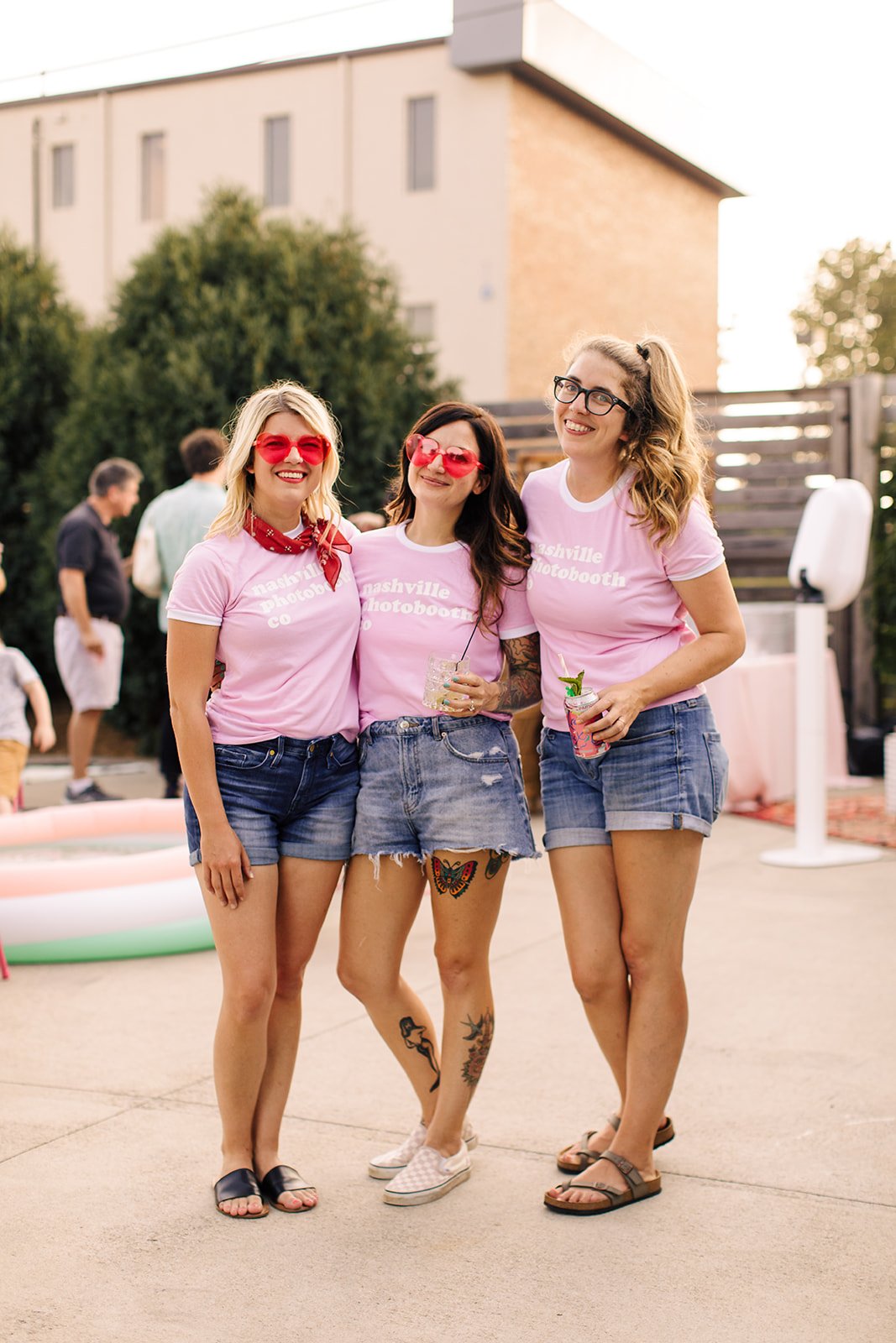 Three women wearing pink Nashville photobooth t-shirts, denim shorts, and sunglasses standing together smiling outdoors at a social gathering, holding drinks.