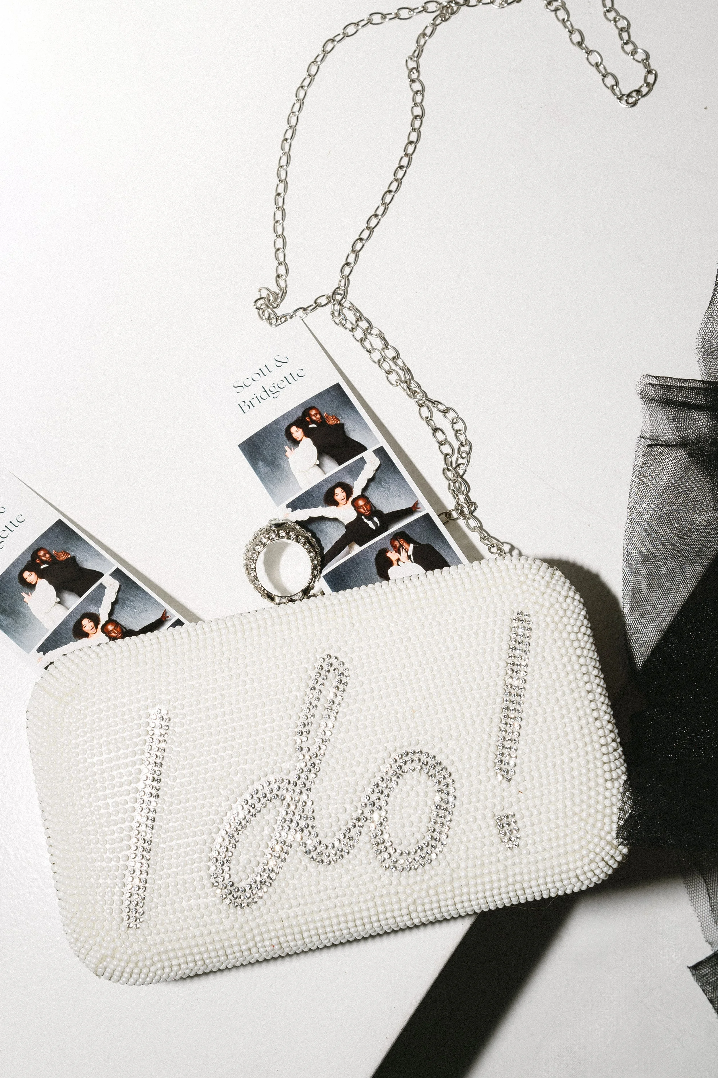 A white beaded clutch with the word "I DO" spelled out in rhinestones, a silver chain strap, a ring attached to the clutch, and two photo booth strips with four photos each, placed on a white surface.