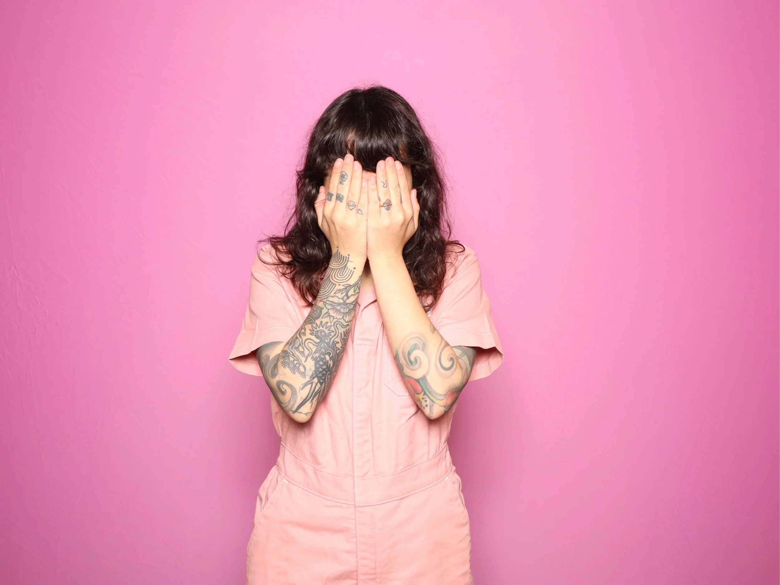 Person in a pink jumpsuit covering face with hands against a pink background.