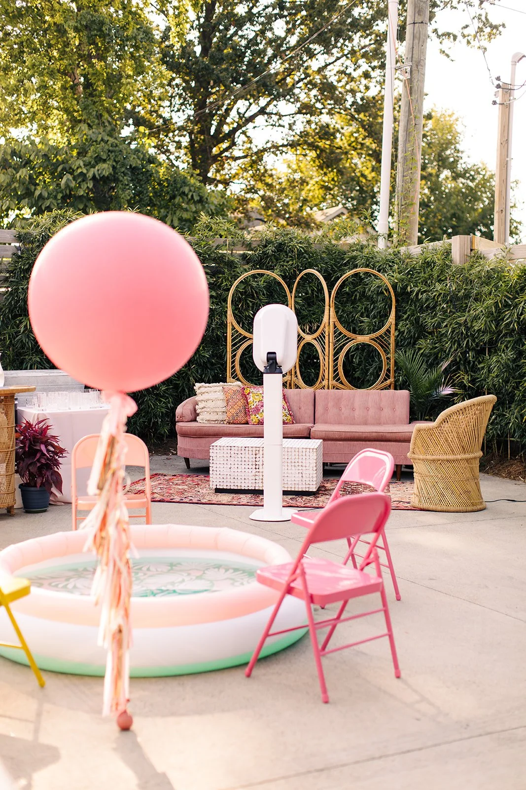 Decorative outdoor patio setup with pink inflatable pool, pink chairs, a pink vintage sofa with pillows, a small white woven table, a gold and wicker chair, lush green foliage, and a backdrop of trees and utility poles.