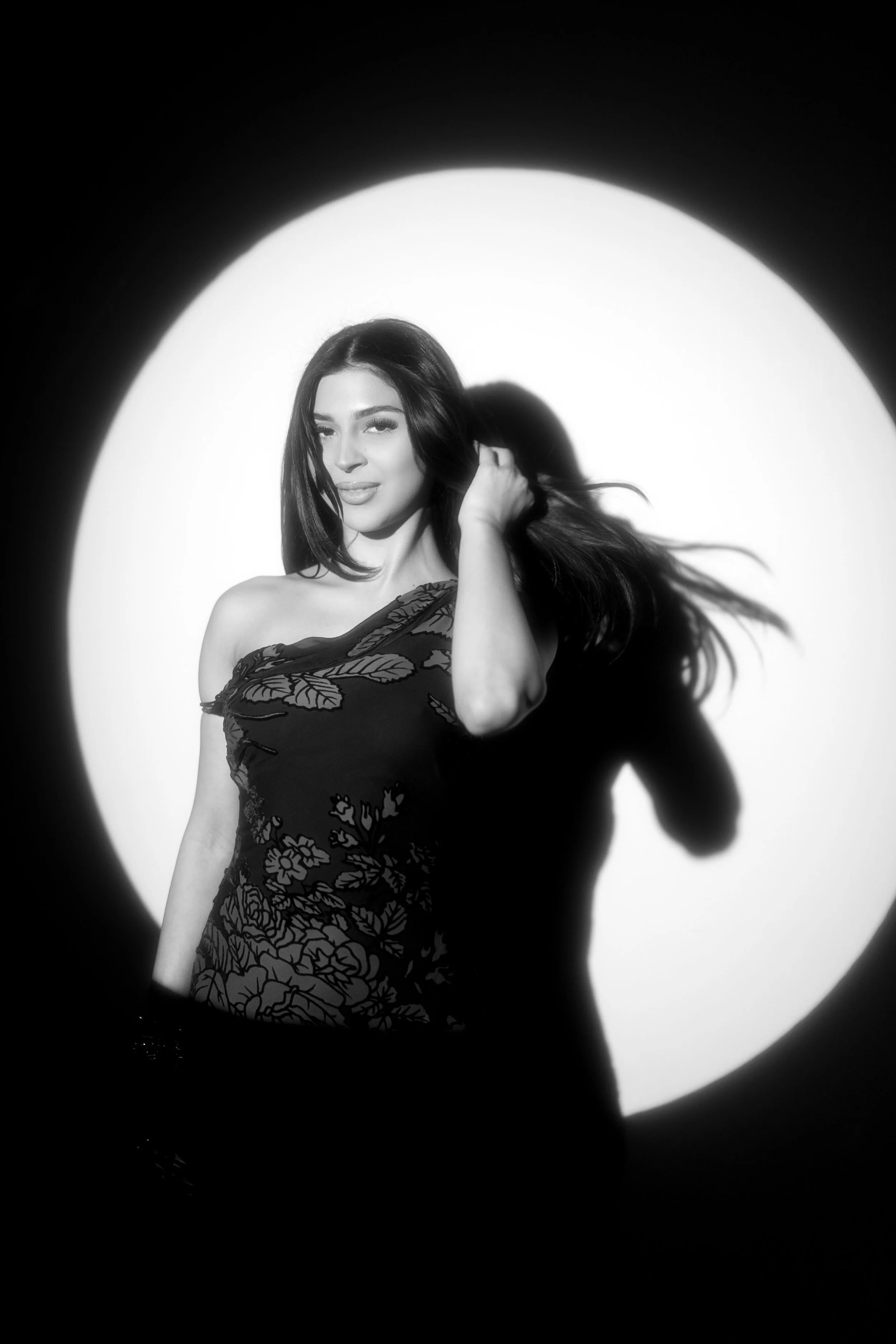Black and white photograph of a woman with long hair, standing in front of a large circular light source, with her hand touching her hair, wearing an off-shoulder dress with floral patterns.