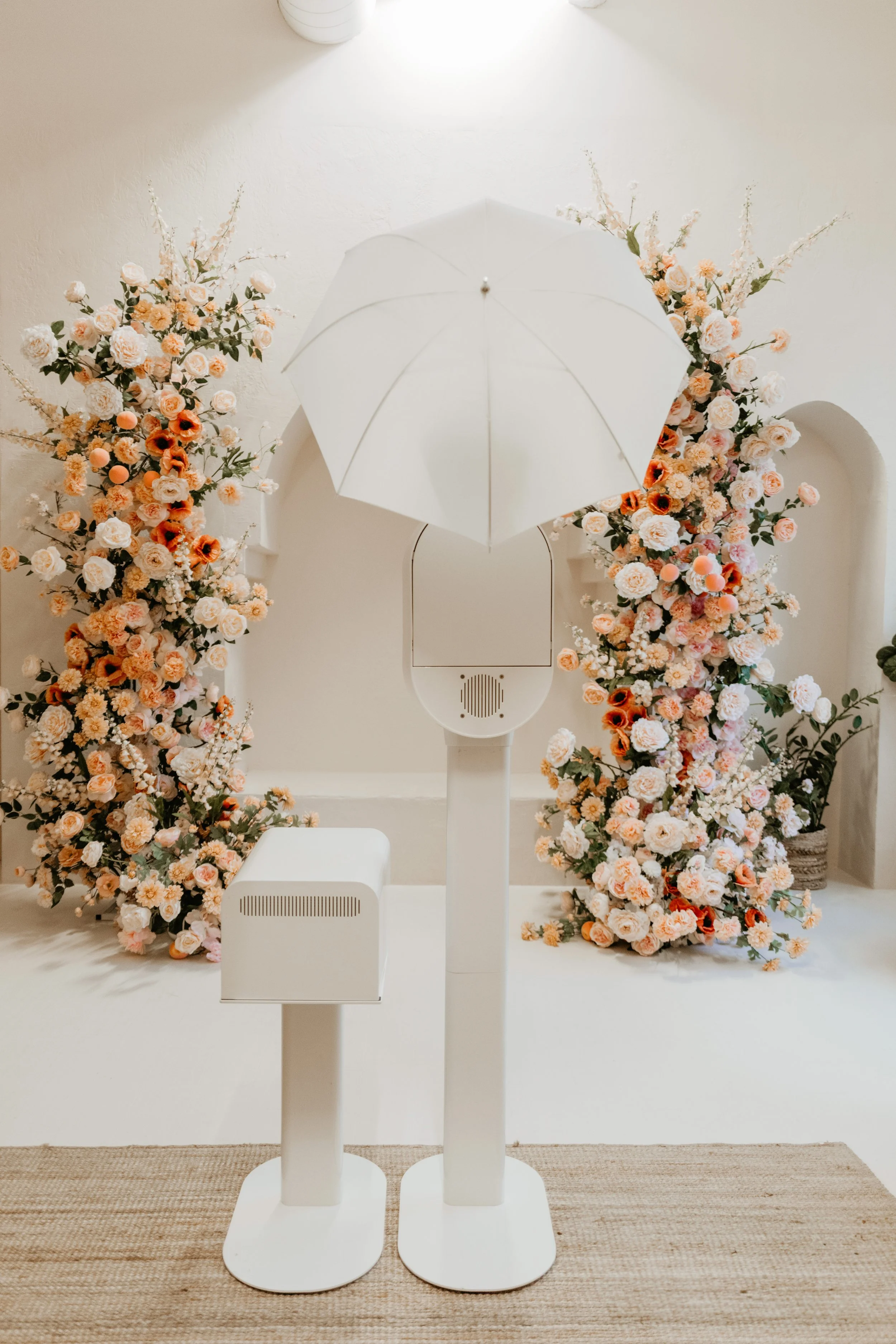 A backdrop of pink and white floral arrangements, with a white umbrella and a white audio device on stands in front.