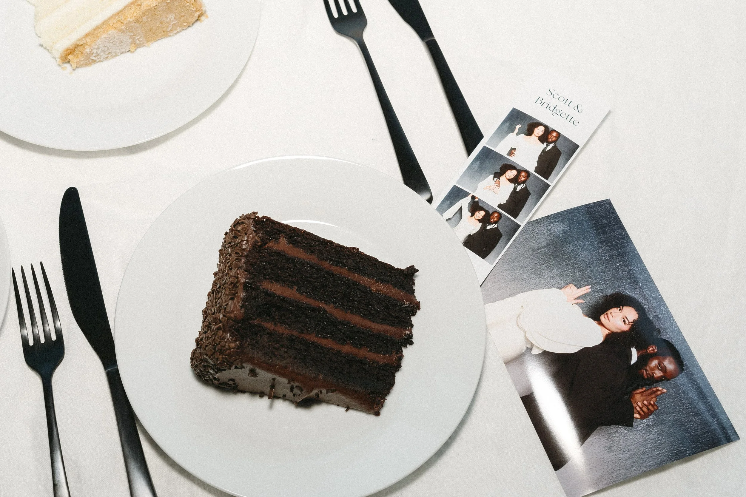 A slice of chocolate cake with multiple layers and chocolate frosting on a white plate, placed on a white table. There are black cutlery pieces beside the plate, a photograph of two women and a man, and a photo of two women leaning against a wall.
