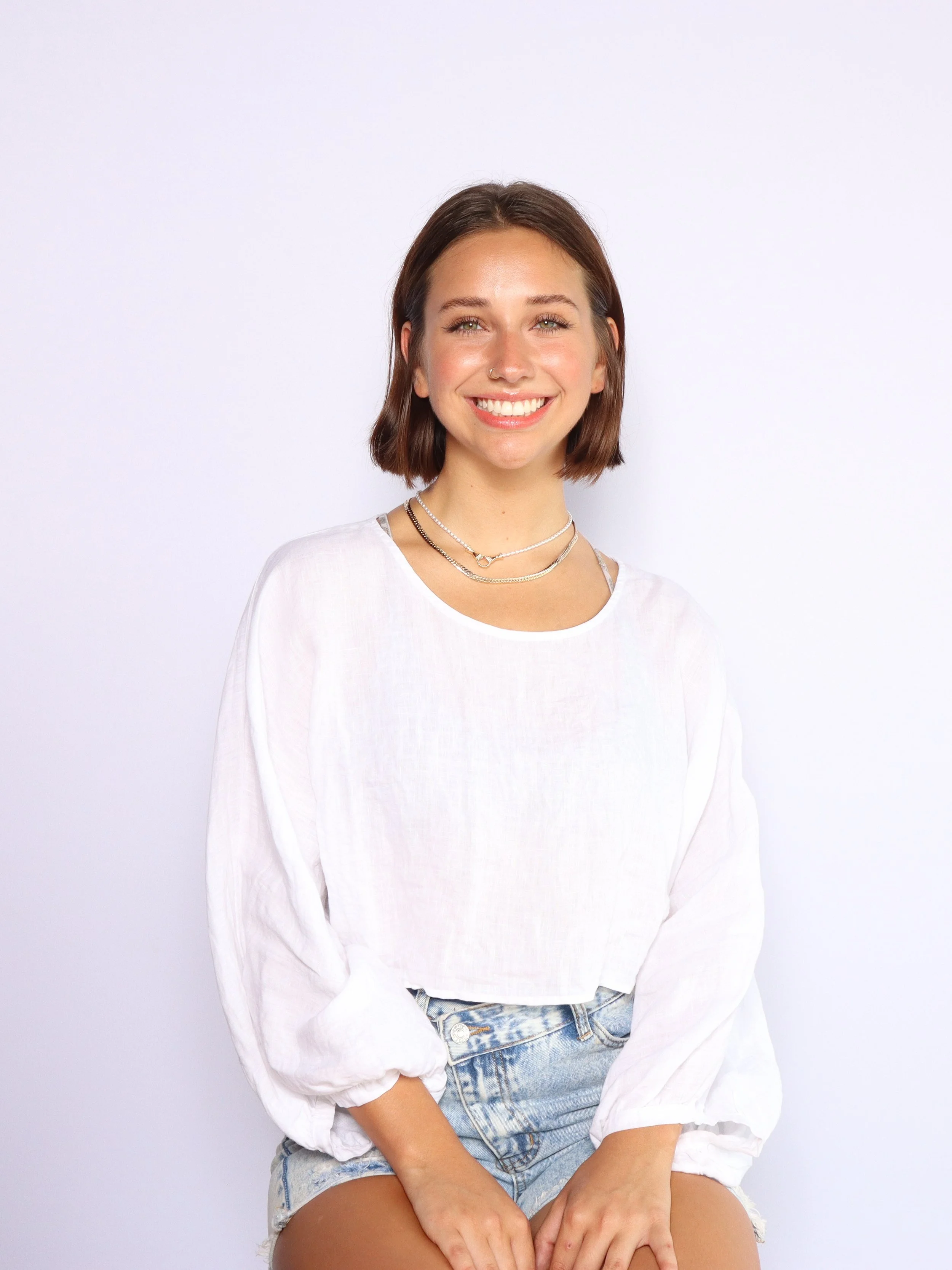 A young woman with shoulder-length brown hair, wearing a white long-sleeve shirt and denim shorts, smiling and sitting against a plain white background.