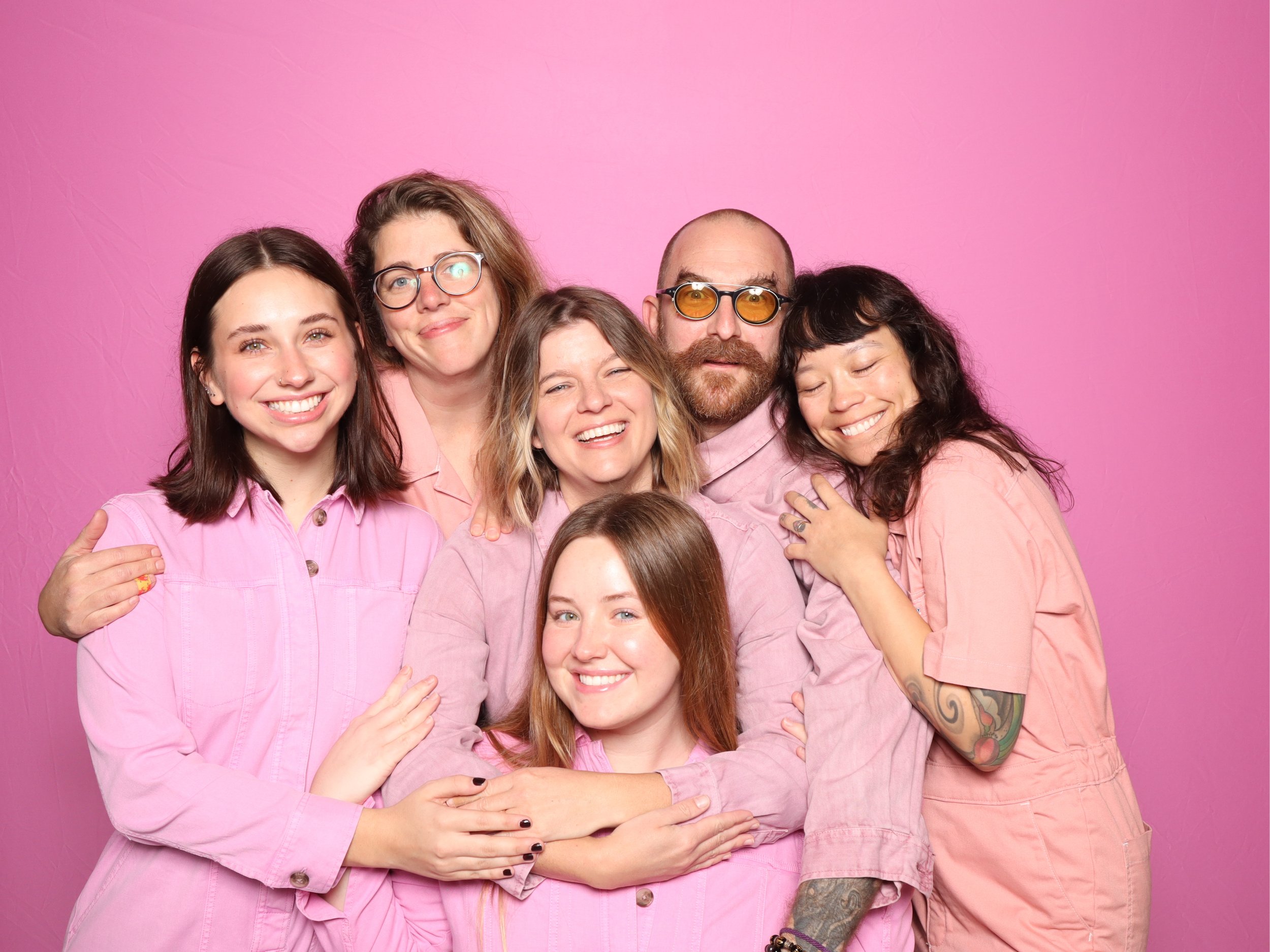 Group of six people hugging in front of a pink background, all wearing pink or light-colored clothing, smiling and celebrating