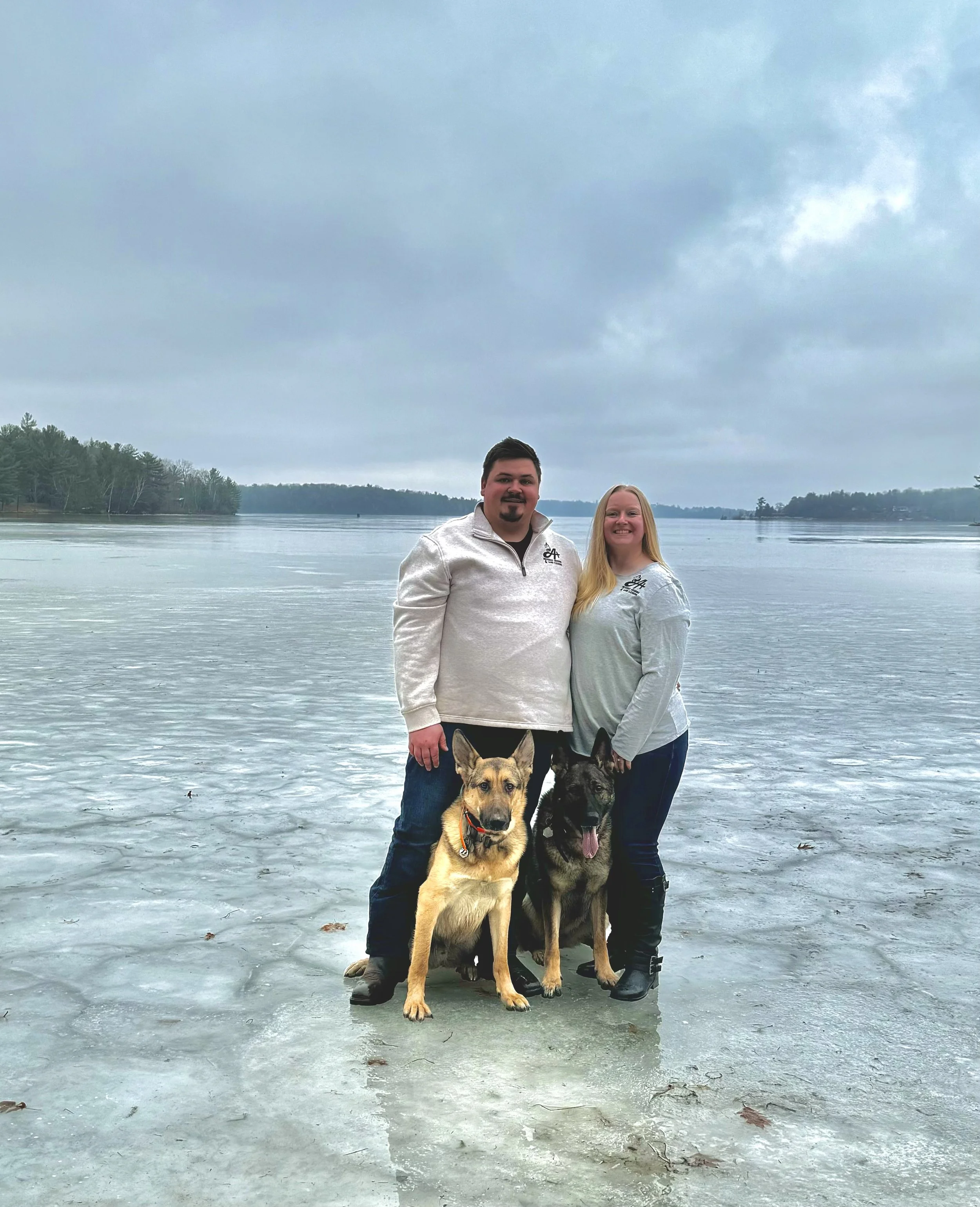 A man and woman standing on a frozen lake with two dogs, a lake and trees in the background.