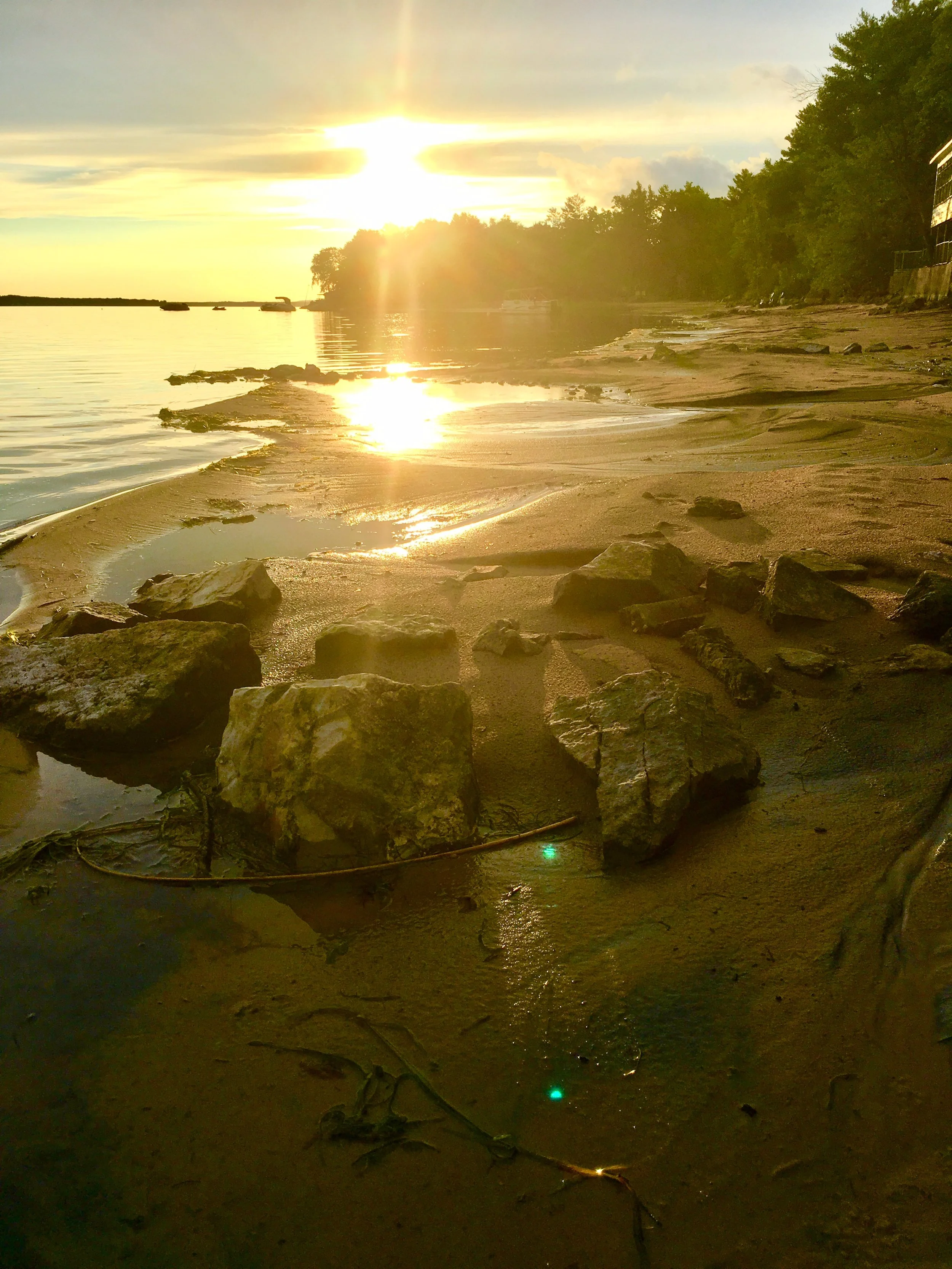 Rocks and dark sand along a shoreline, with sunlight reflected on the water.