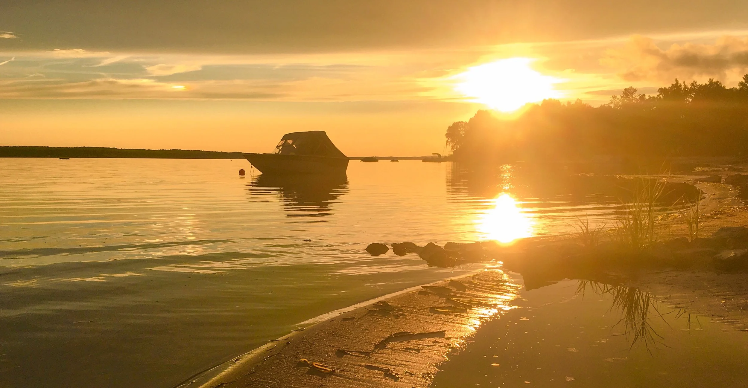An anchored boat and its reflection in still water beside a sunlit shore.