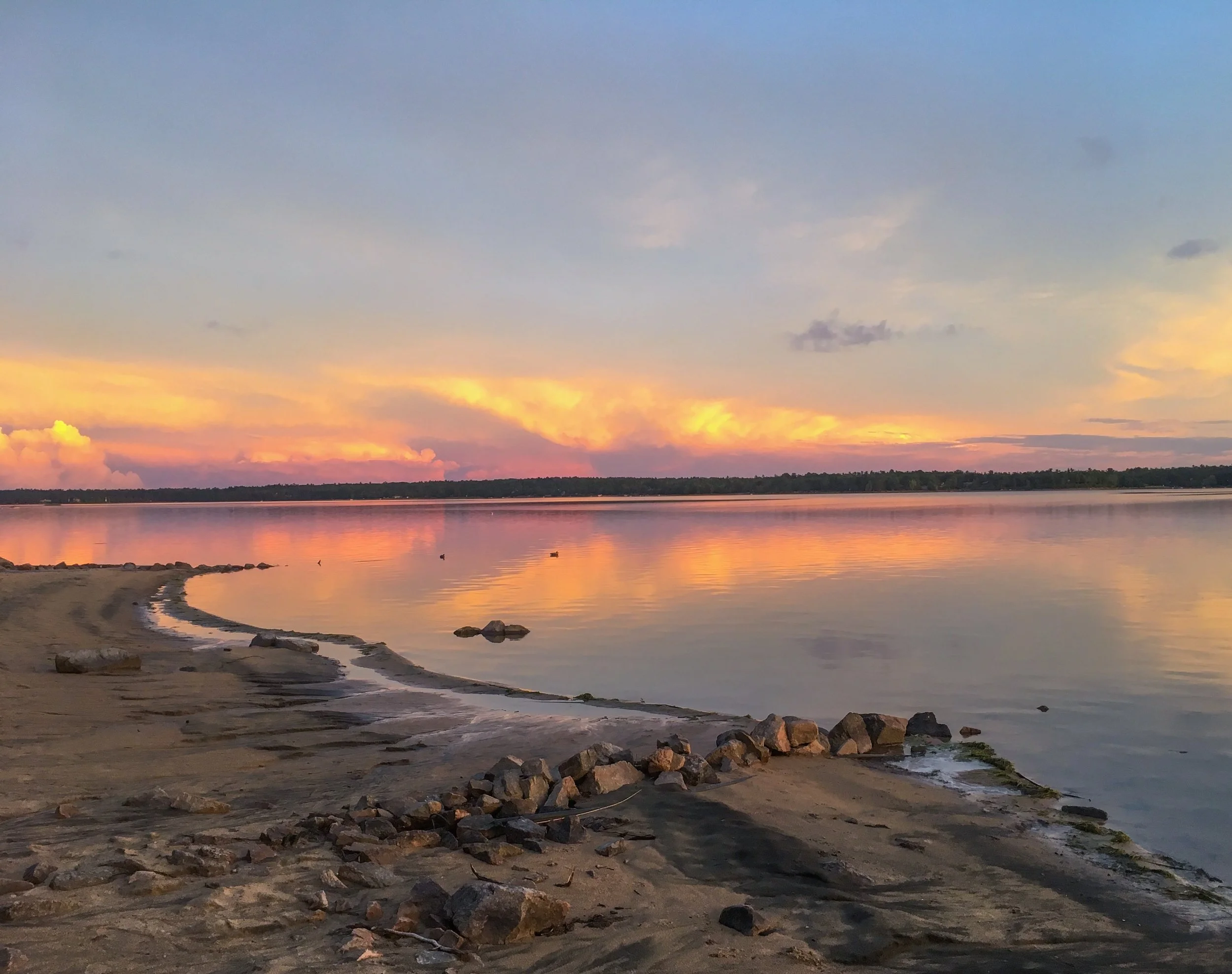 Quiet lake water, a curving sandy shoreline and an open sky in soft evening light.