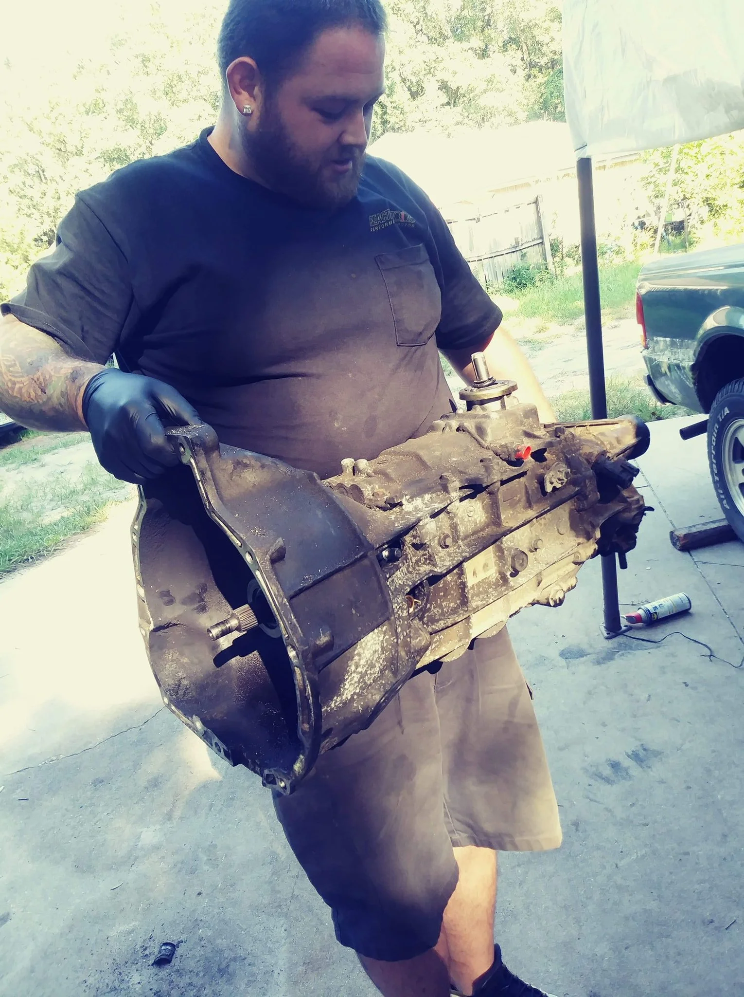 A man wearing a black shirt with a logo, black gloves, and shorts, holding a used car transmission outdoors.