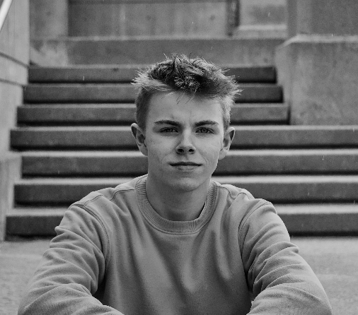 Black and white photo of a young man with short, messy hair, sitting on outdoor stairs, looking at the camera with a neutral expression.