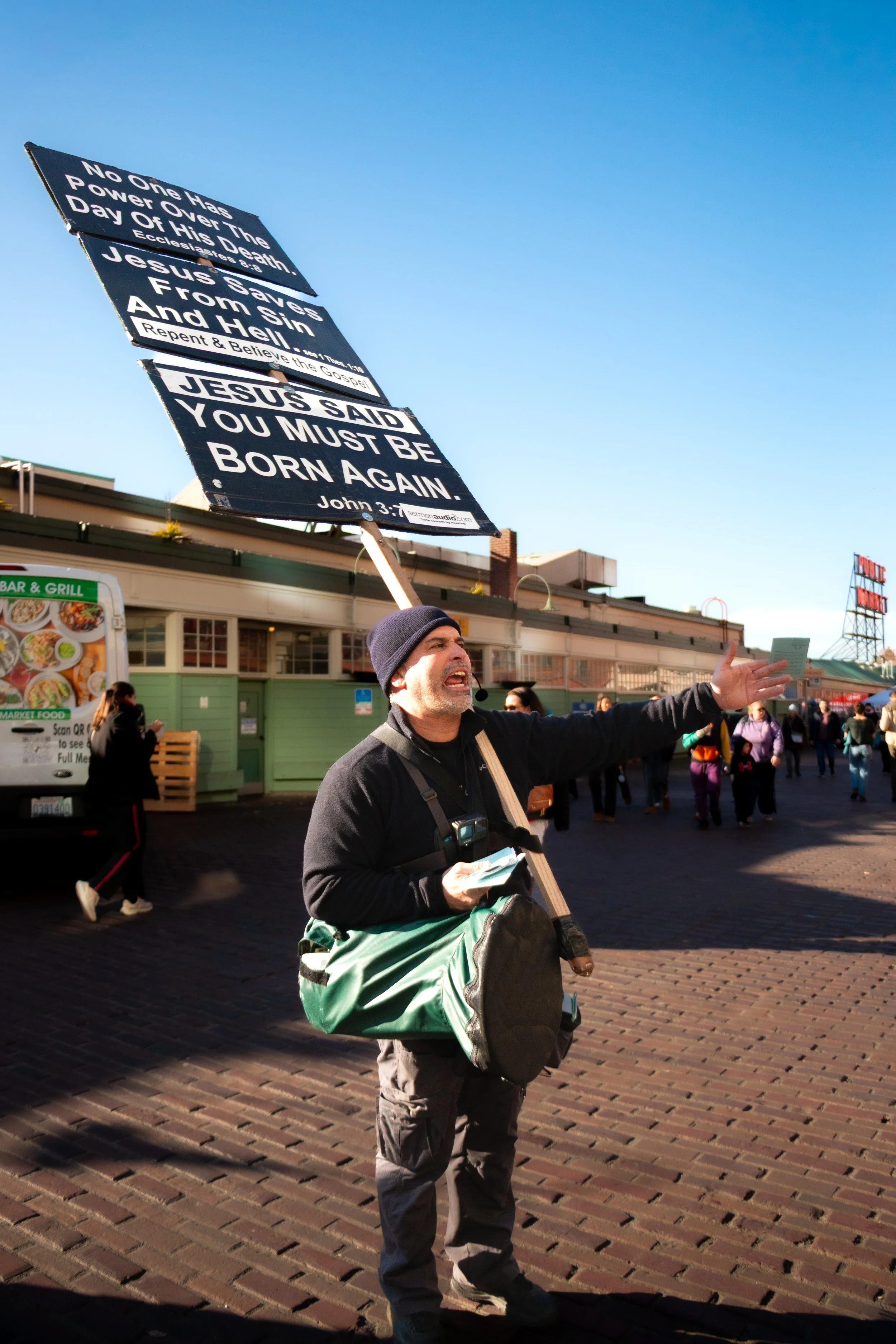 A man speaking passionately and gesturing outdoors at a public gathering, holding a sign with religious messages and scripture.