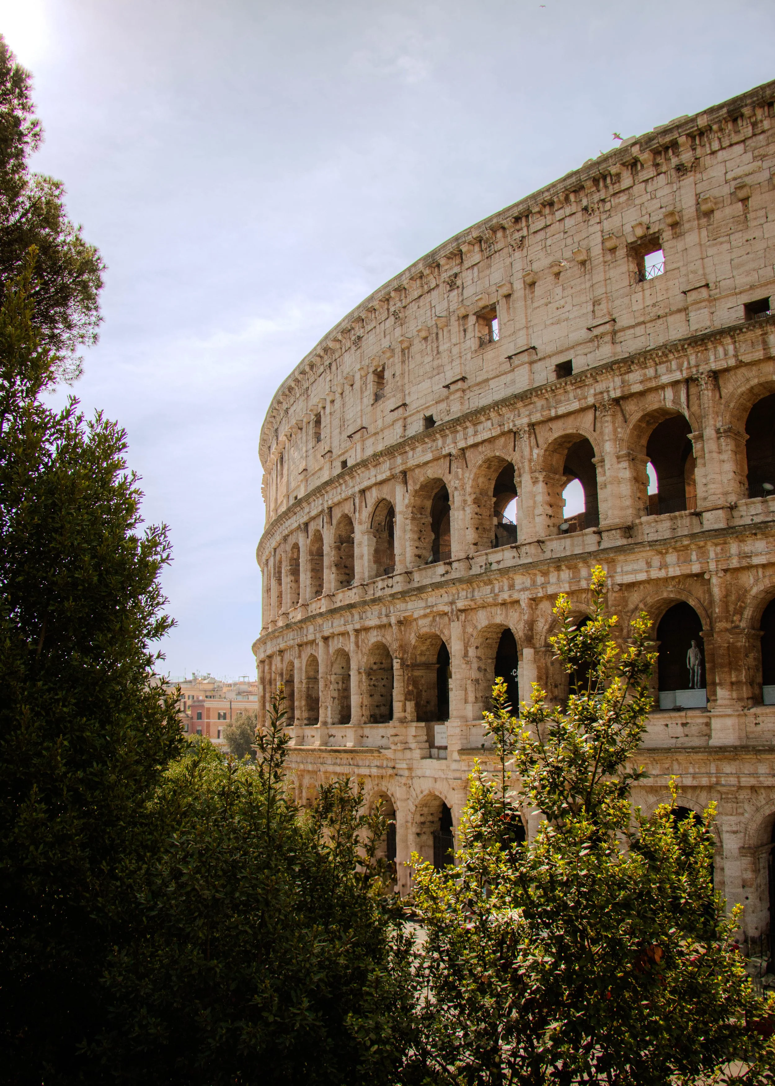 Exterior view of the Colosseum in Rome, Italy, with greenery in the foreground and a partly cloudy sky.