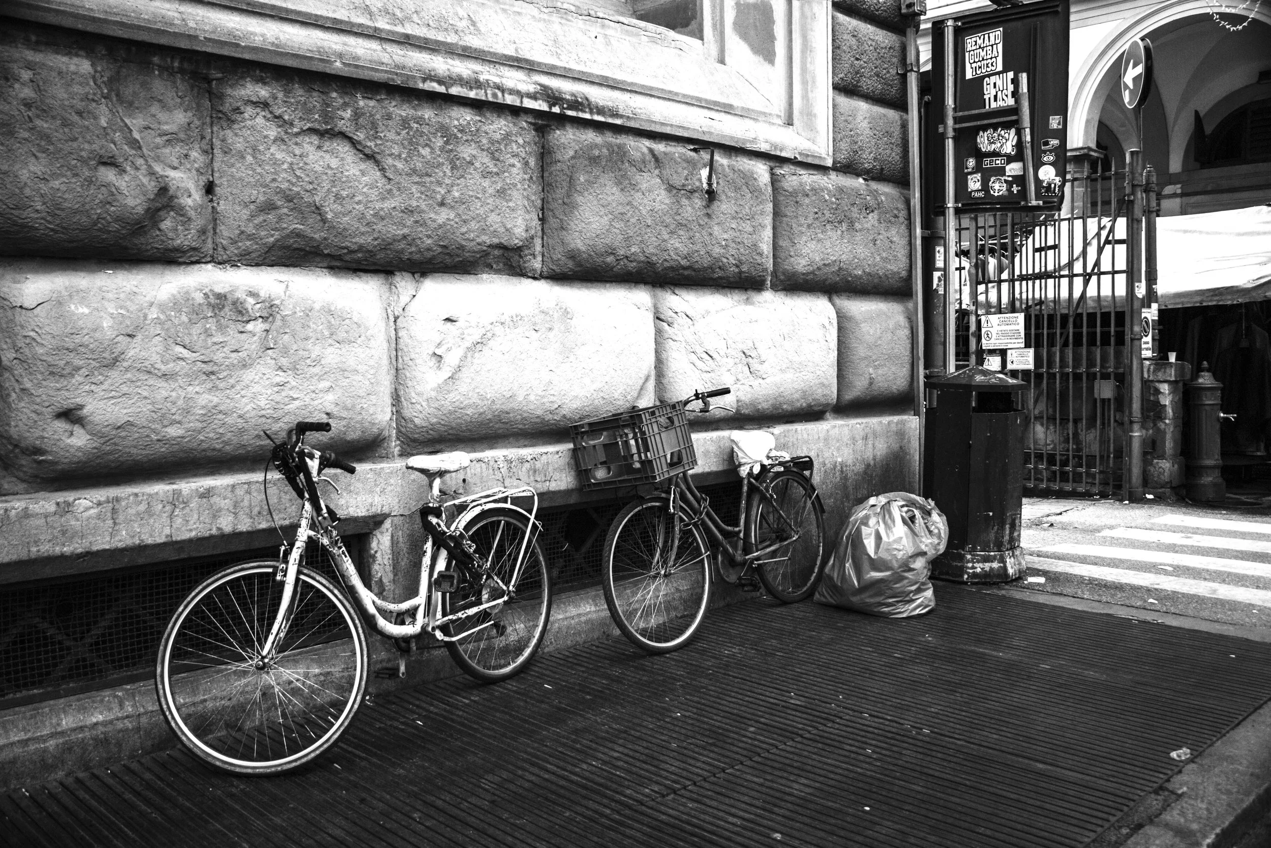 Two bicycles leaning against a stone wall, with a garbage bag beside them near a metal gate and trash can on a city street.