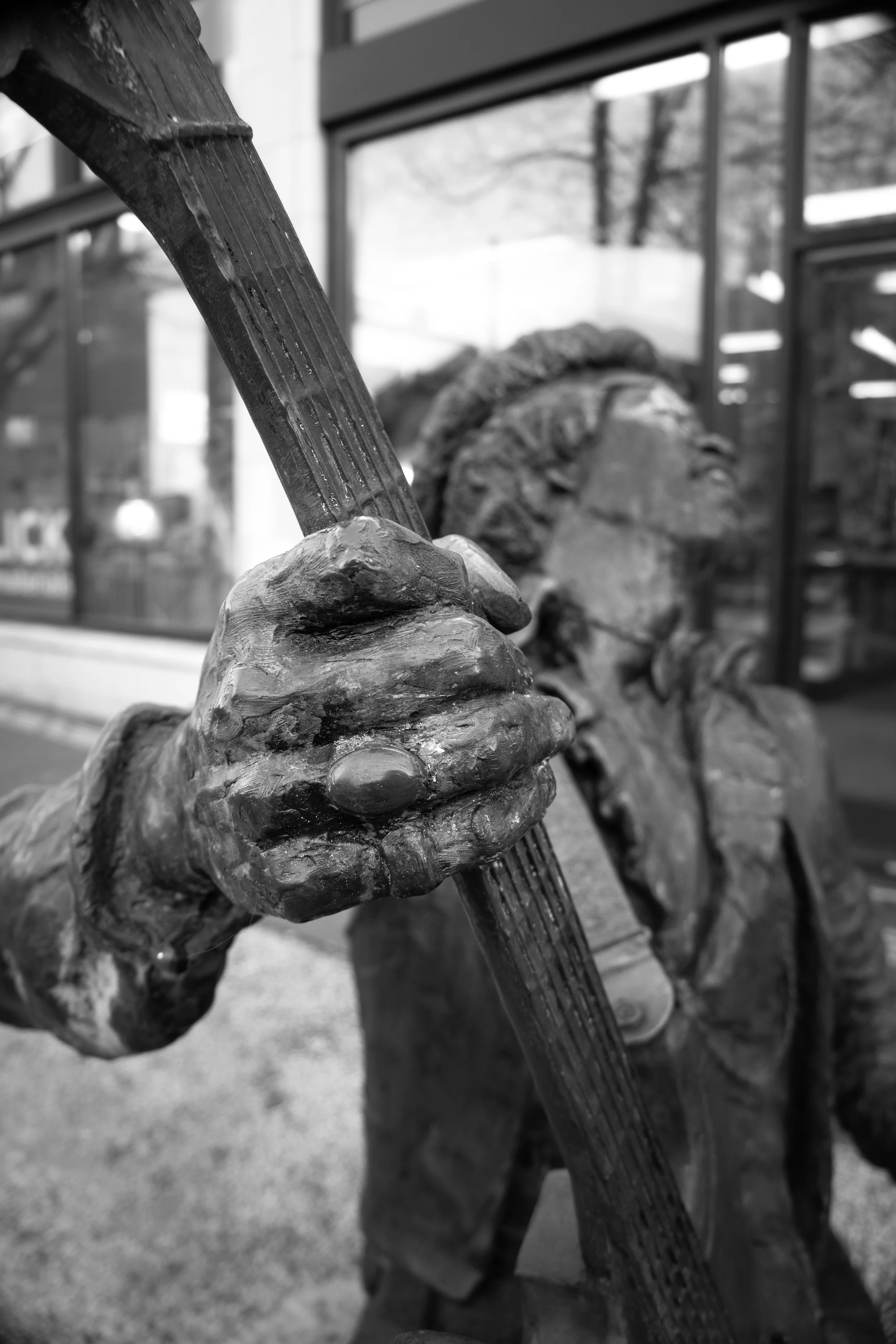 Close-up of a bronze statue of a person holding a musical instrument, with a blurred background of a building and trees.