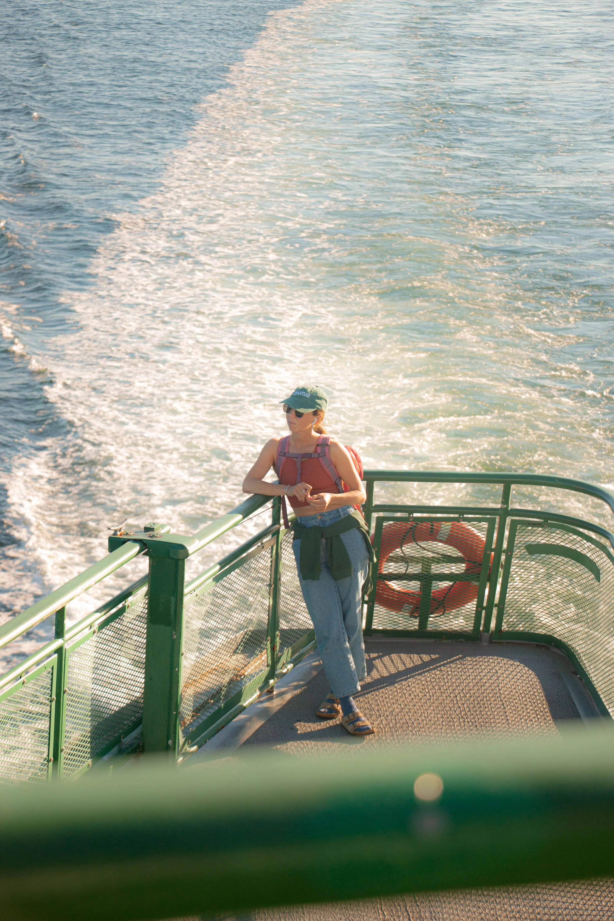 A woman standing on the deck of a ferry, looking out at the water, wearing sunglasses, a cap, a red tank top, jeans, and sandals, with a backpack, during sunset.
