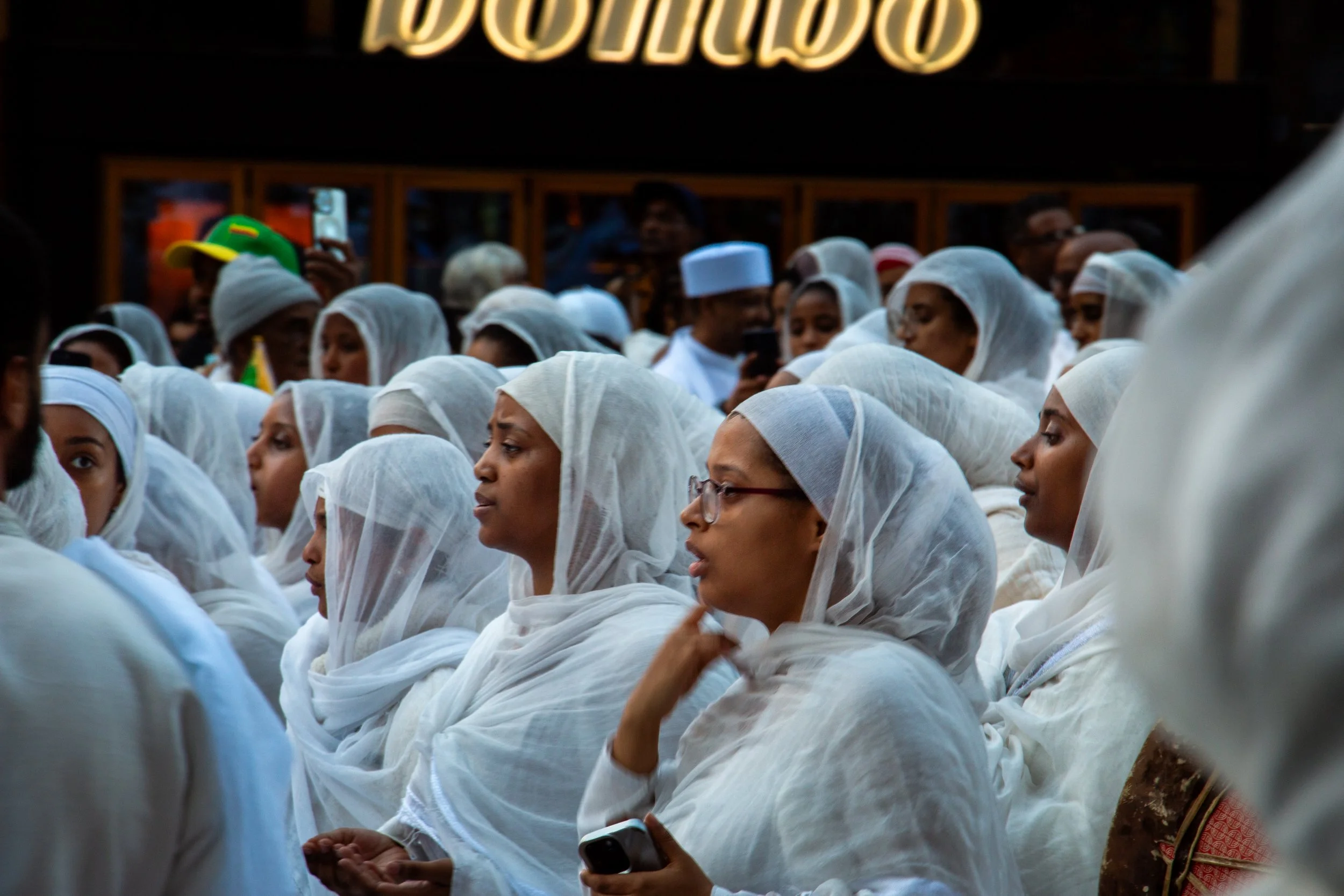 Group of women wearing white traditional attire with head coverings, sitting silently, some holding phones, in an outdoor setting during daytime.