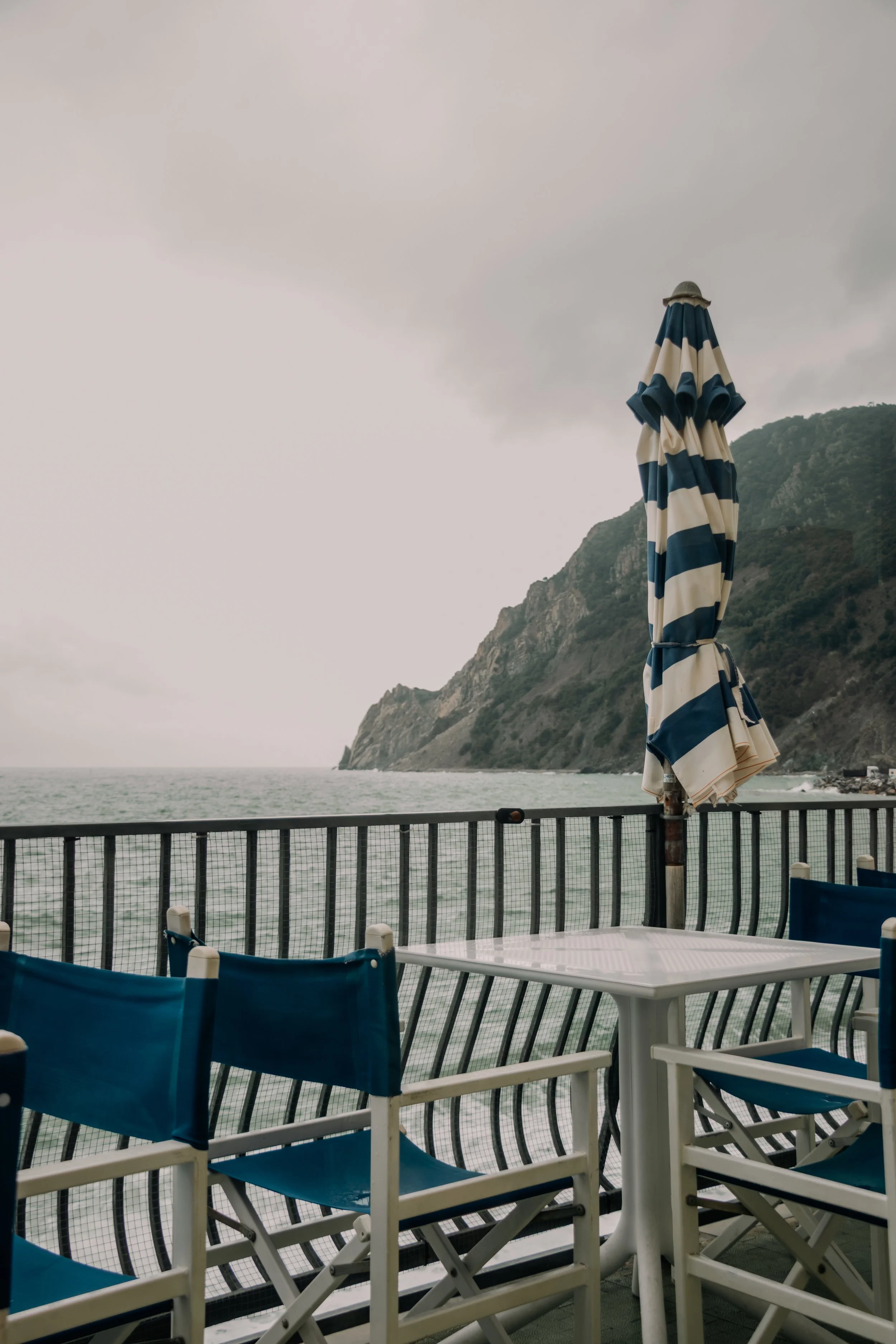 Empty outdoor patio with white and blue chairs, a white table, and a striped blue and white umbrella overlooking a sea and mountainous coastline under cloudy sky.