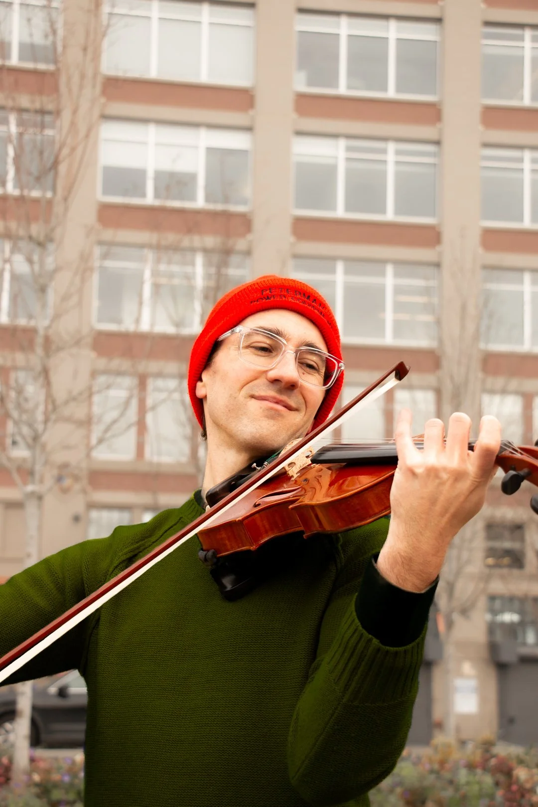 A man wearing glasses and a red beanie playing a violin outdoors in front of a multi-story apartment building.