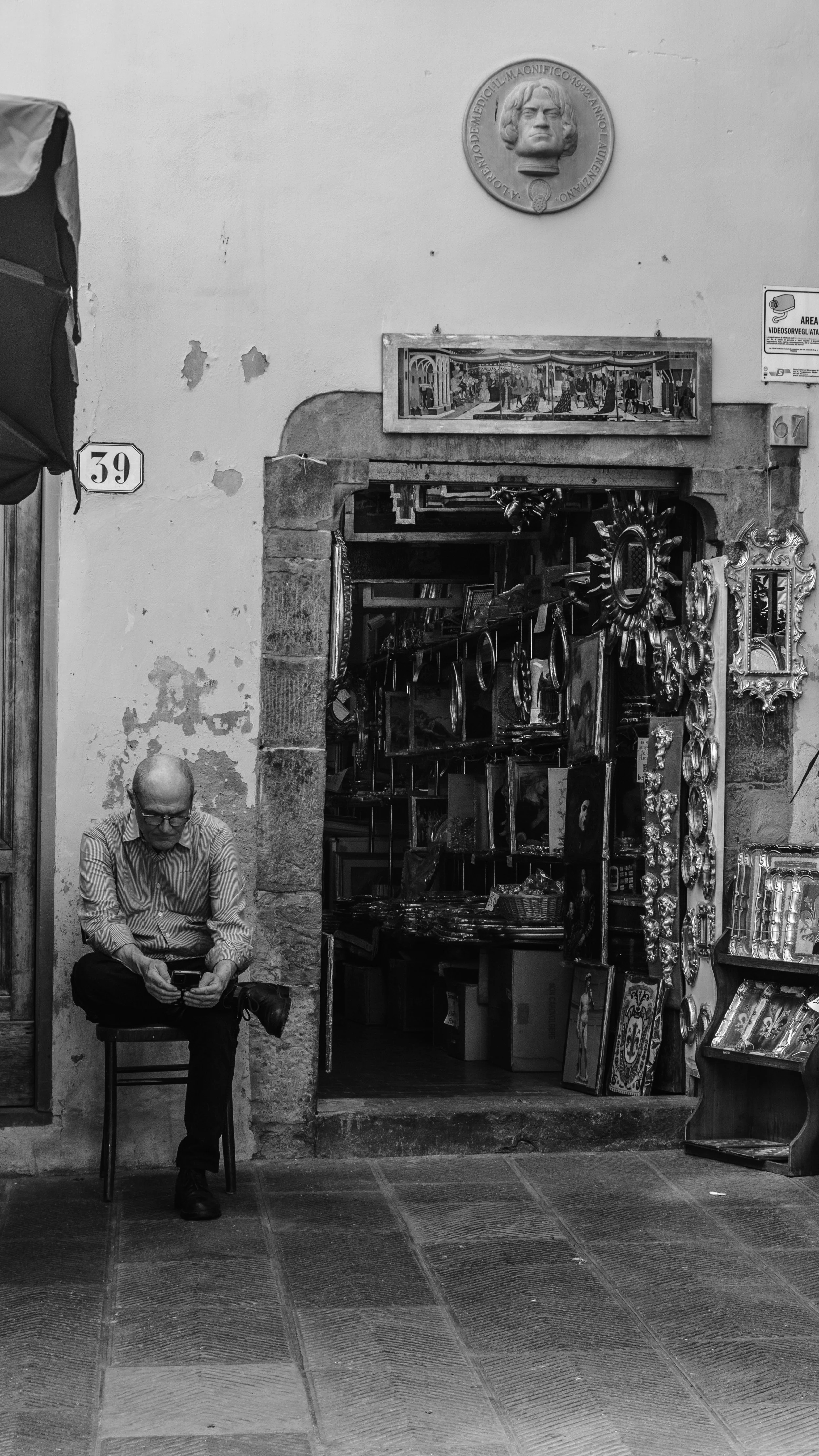 A man sitting on a small chair outside a shop, looking at his phone, in a street scene.