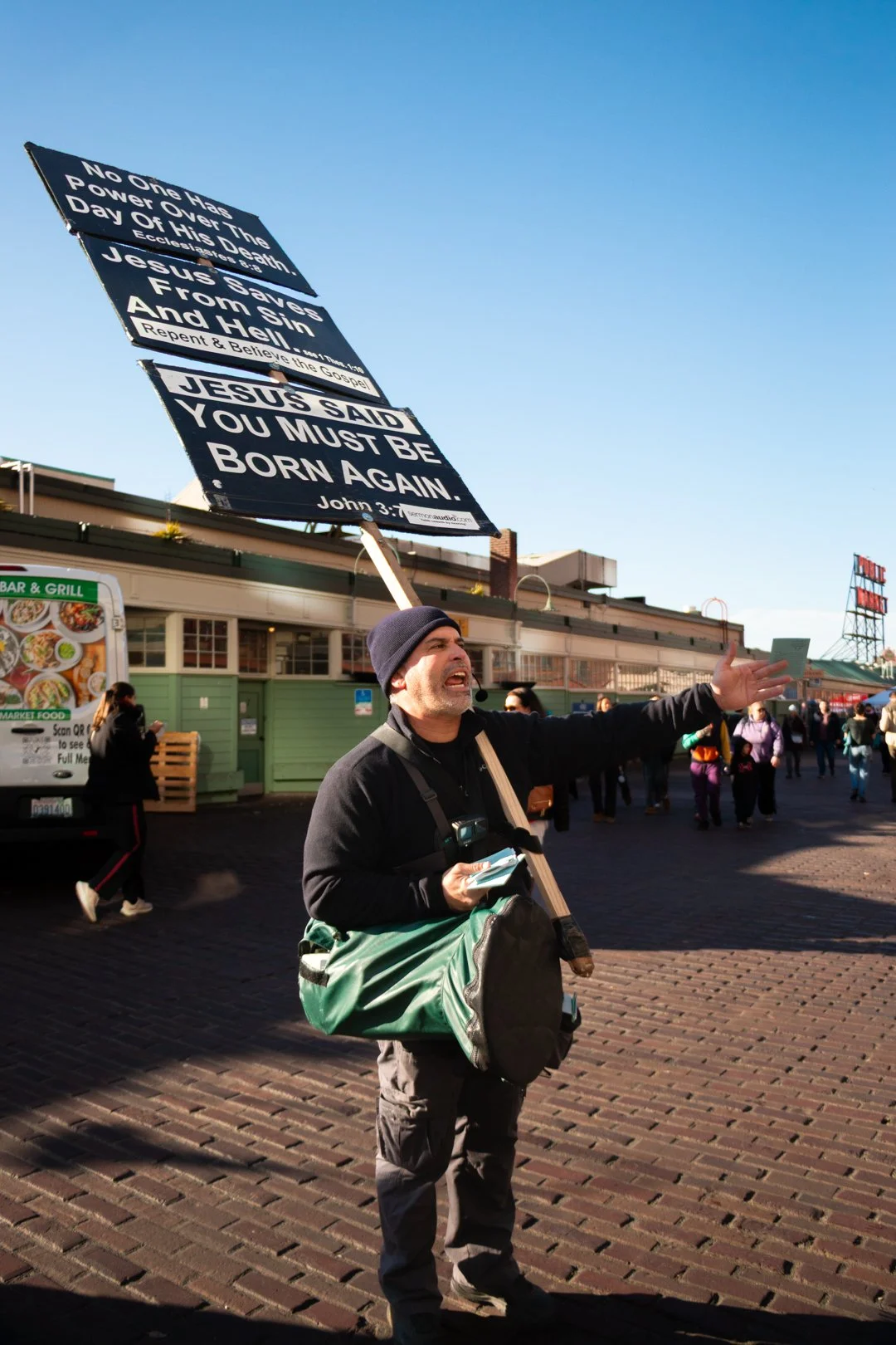 A street preacher holding a sign with a biblical quote, gesturing outward with a crowd in the background.