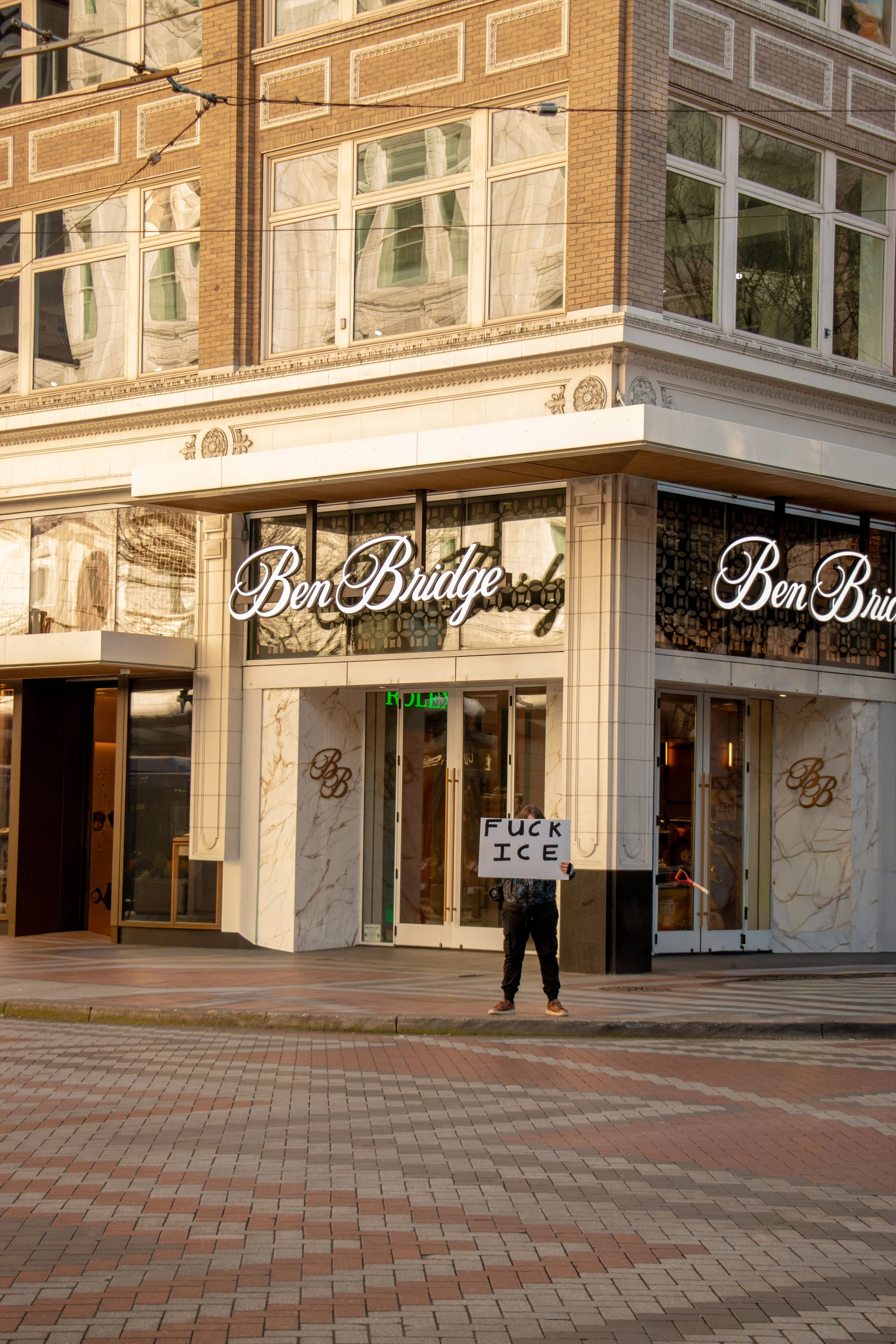 A person standing outside a building holding a sign that says 'FUCK ICE.' The building has a sign that reads 'Ben Bridge' and appears to be a jewelry store, with marble accents and large windows.