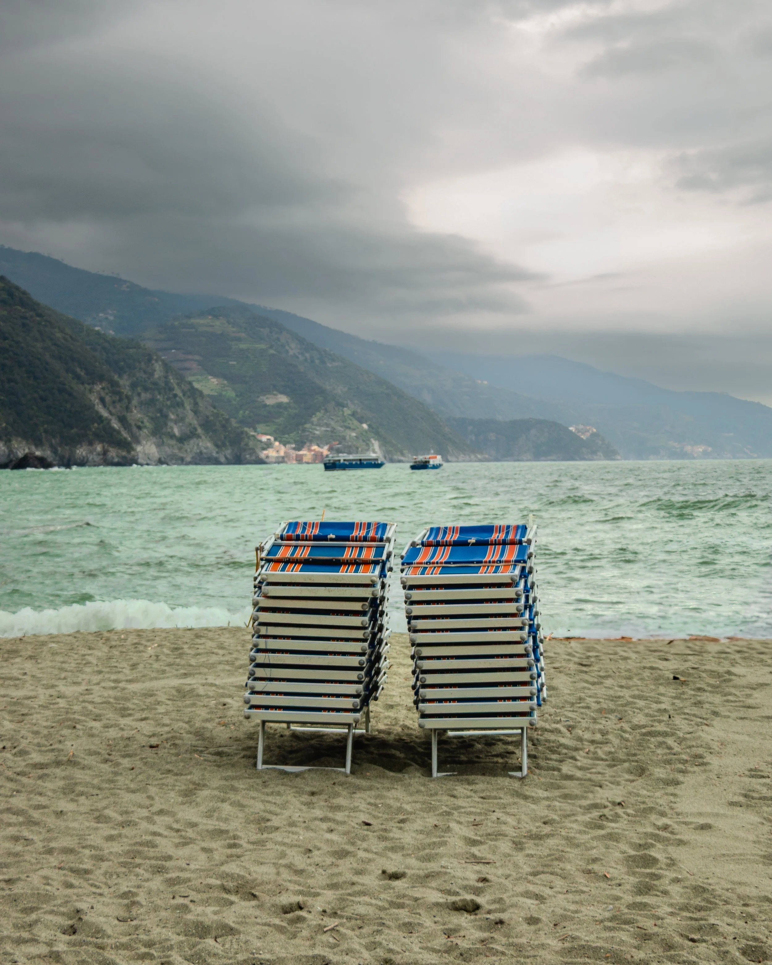 Empty lounge chairs on a sandy beach facing the ocean, with mountains and boats in the background under cloudy skies.