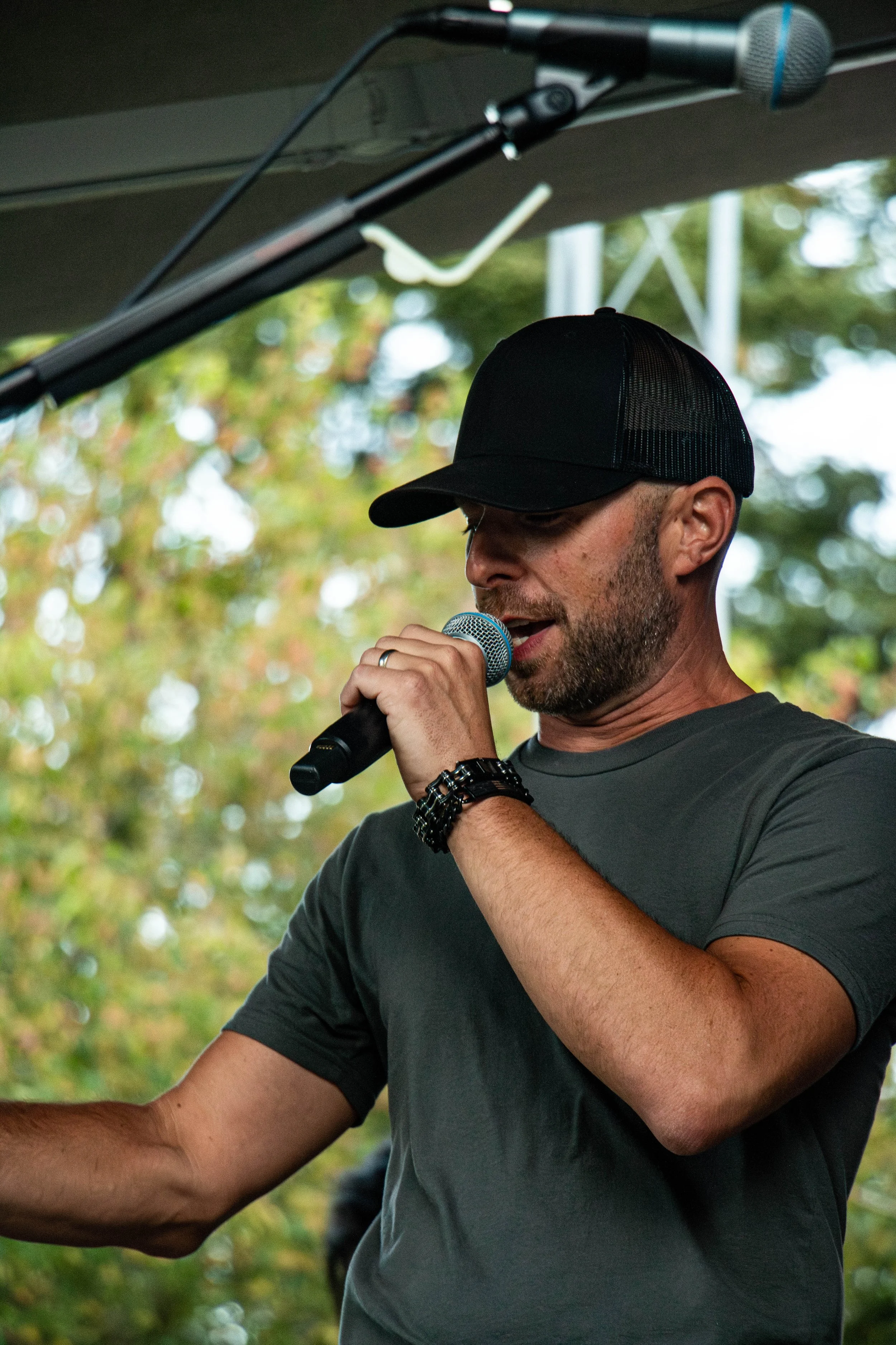 A man wearing a black baseball cap and a gray t-shirt speaks into a microphone at an outdoor event, with a background of trees and cloudy sky.