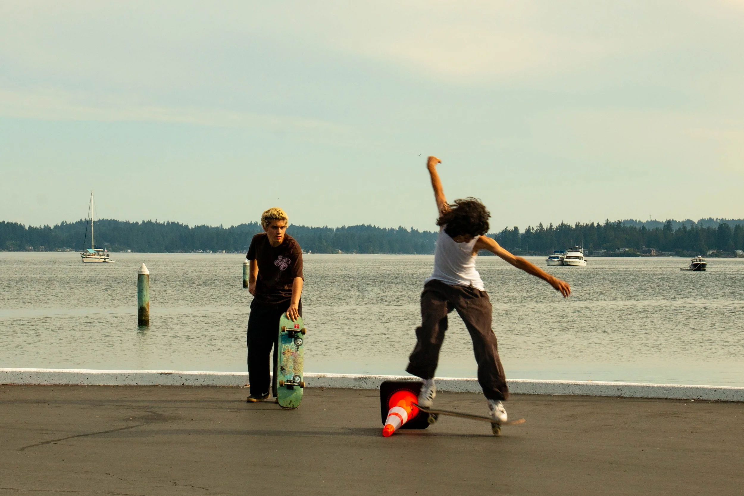 Two skateboarders near a waterfront with boats. One skateboarder is doing a trick over a traffic cone while the other watches.