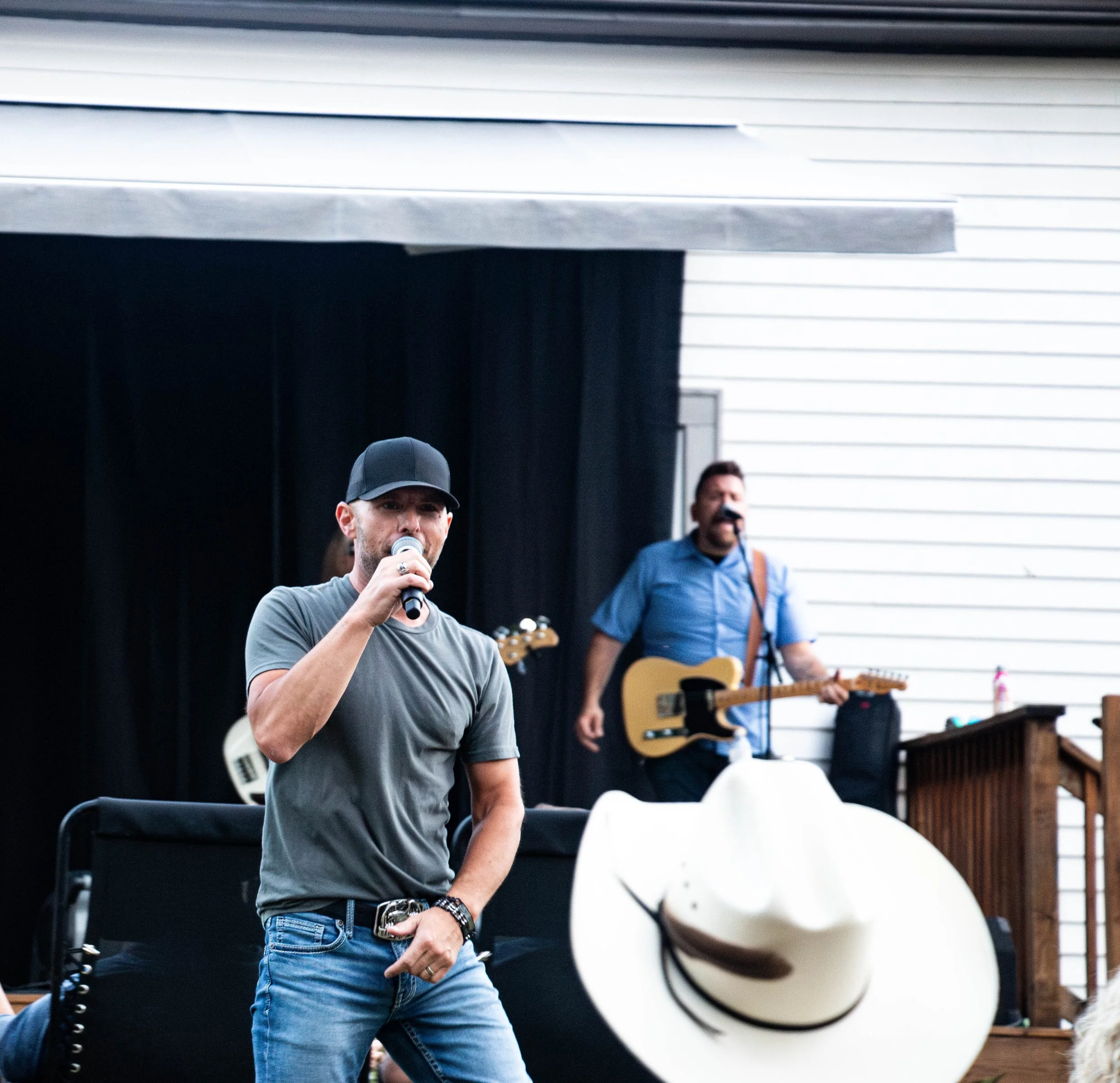 Man in a gray t-shirt and black baseball cap holding a microphone on an outdoor stage, with a musician playing guitar in the background and a white cowboy hat in the foreground.