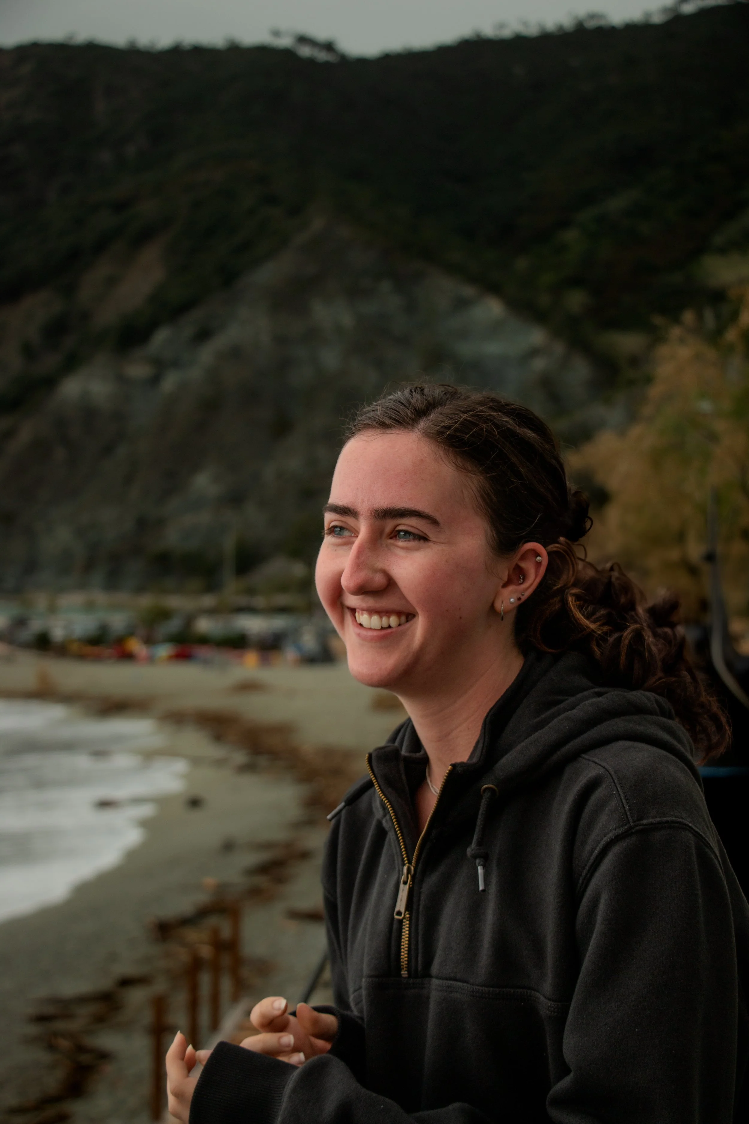 A young woman with dark brown hair in a ponytail, smiling, standing by the beach with mountains in the background.