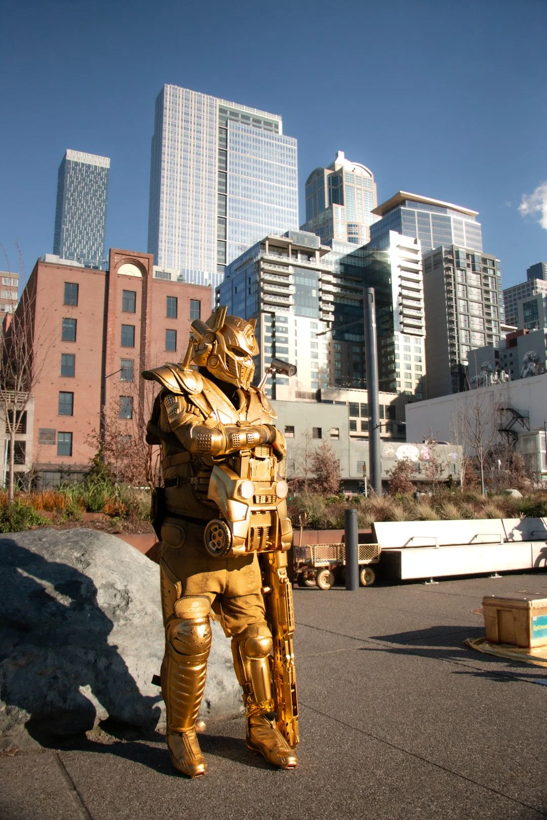 A person in a full-body gold costume resembling a robot or sci-fi character standing outside in an urban area with tall buildings in the background.