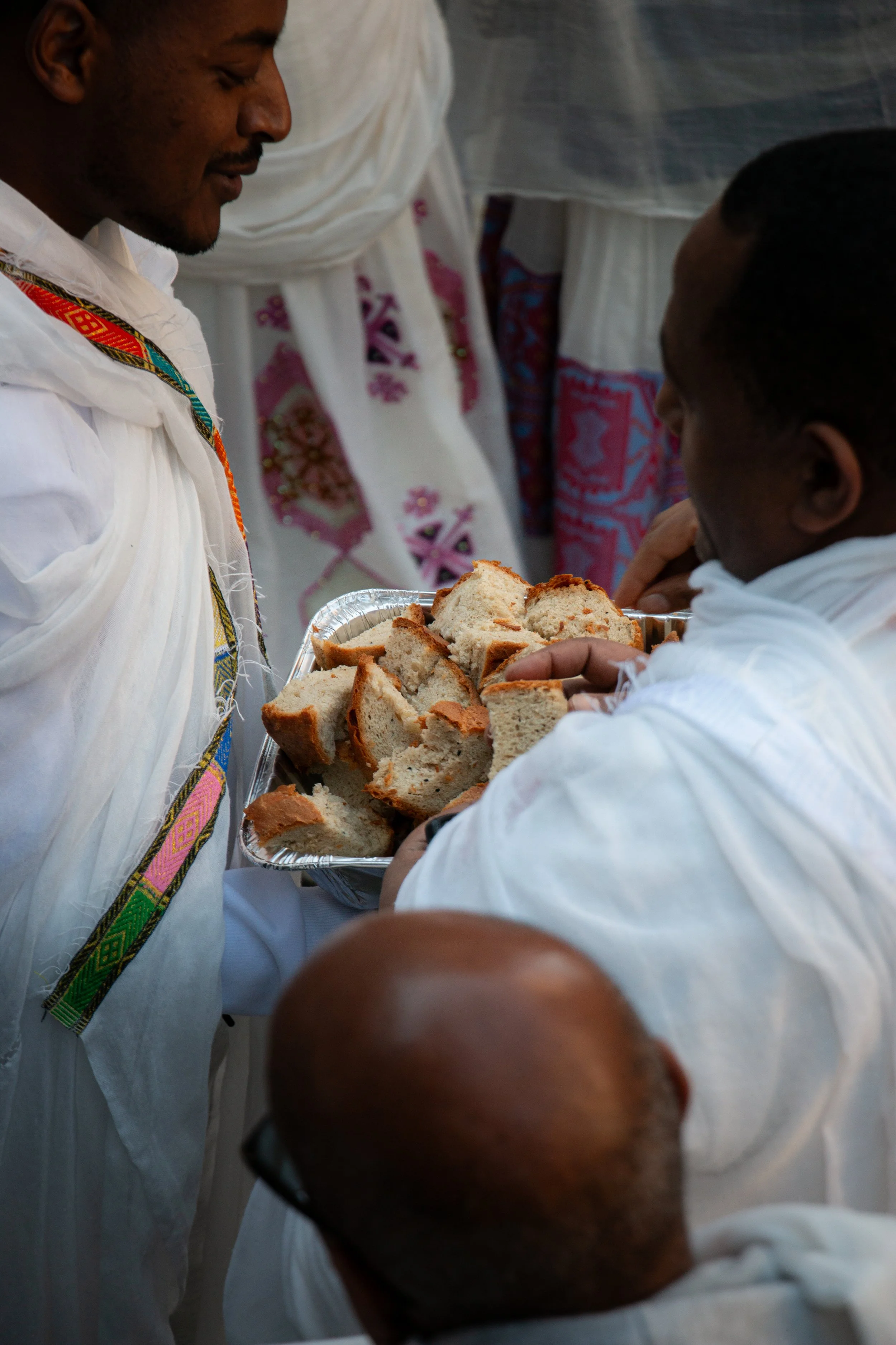 Two men in traditional white clothing sharing slices of bread, with one man offering the bread on a tray to the other.