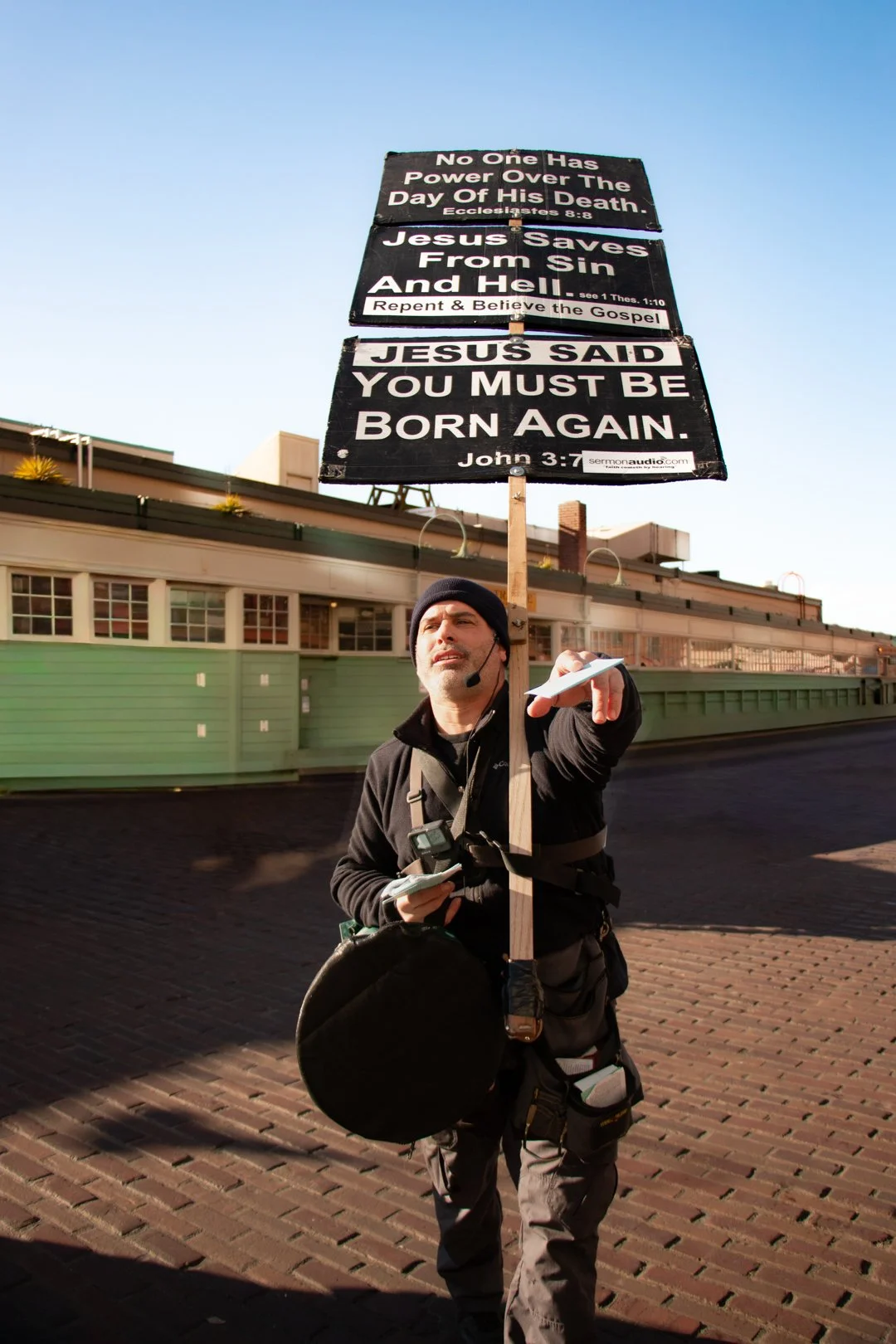 Man standing on brick pavement holding a tall sign with religious messages, pointing with his right hand, wearing a black jacket and a beanie.