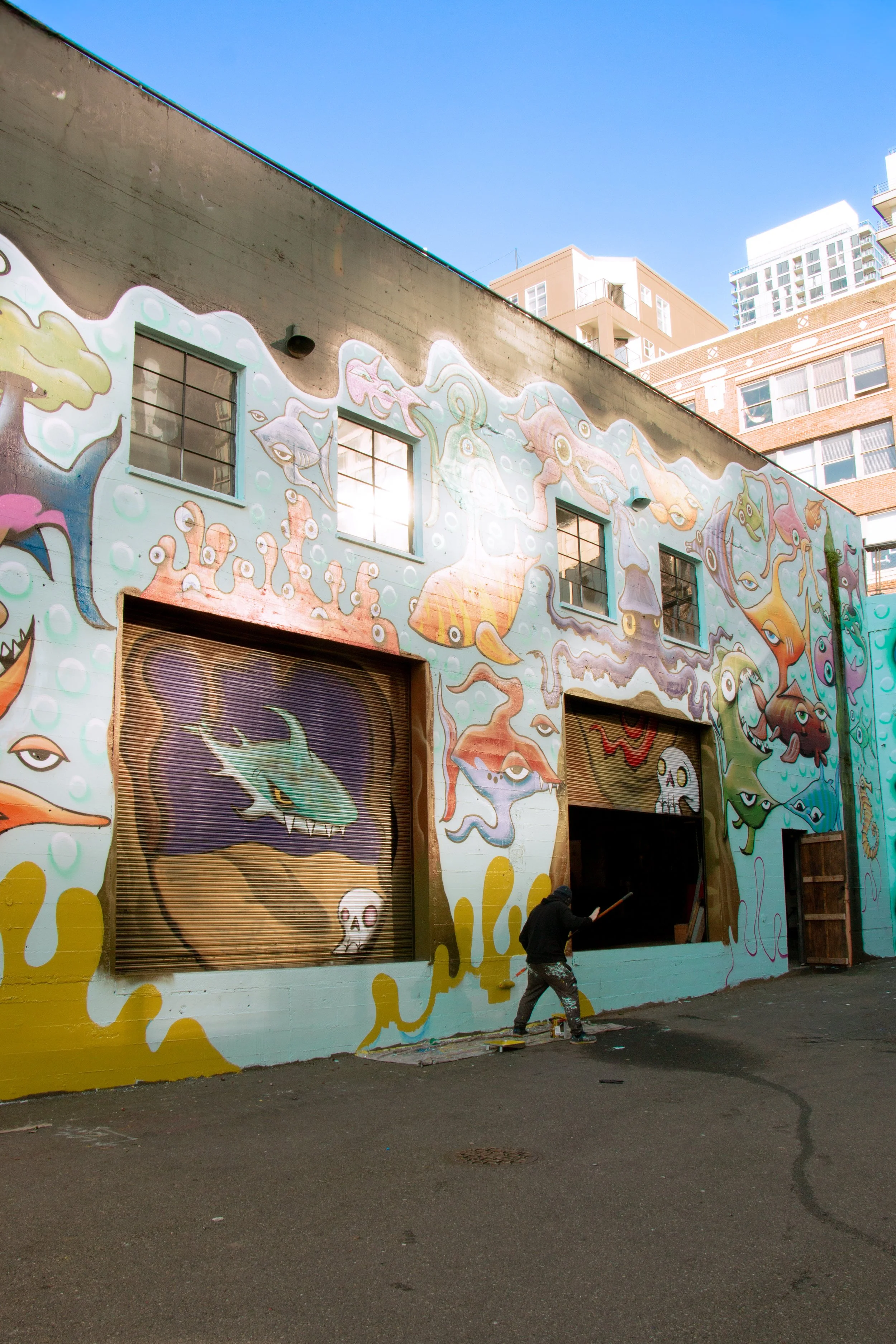 Street artist painting a colorful mural with underwater creatures and abstract designs on a building wall.