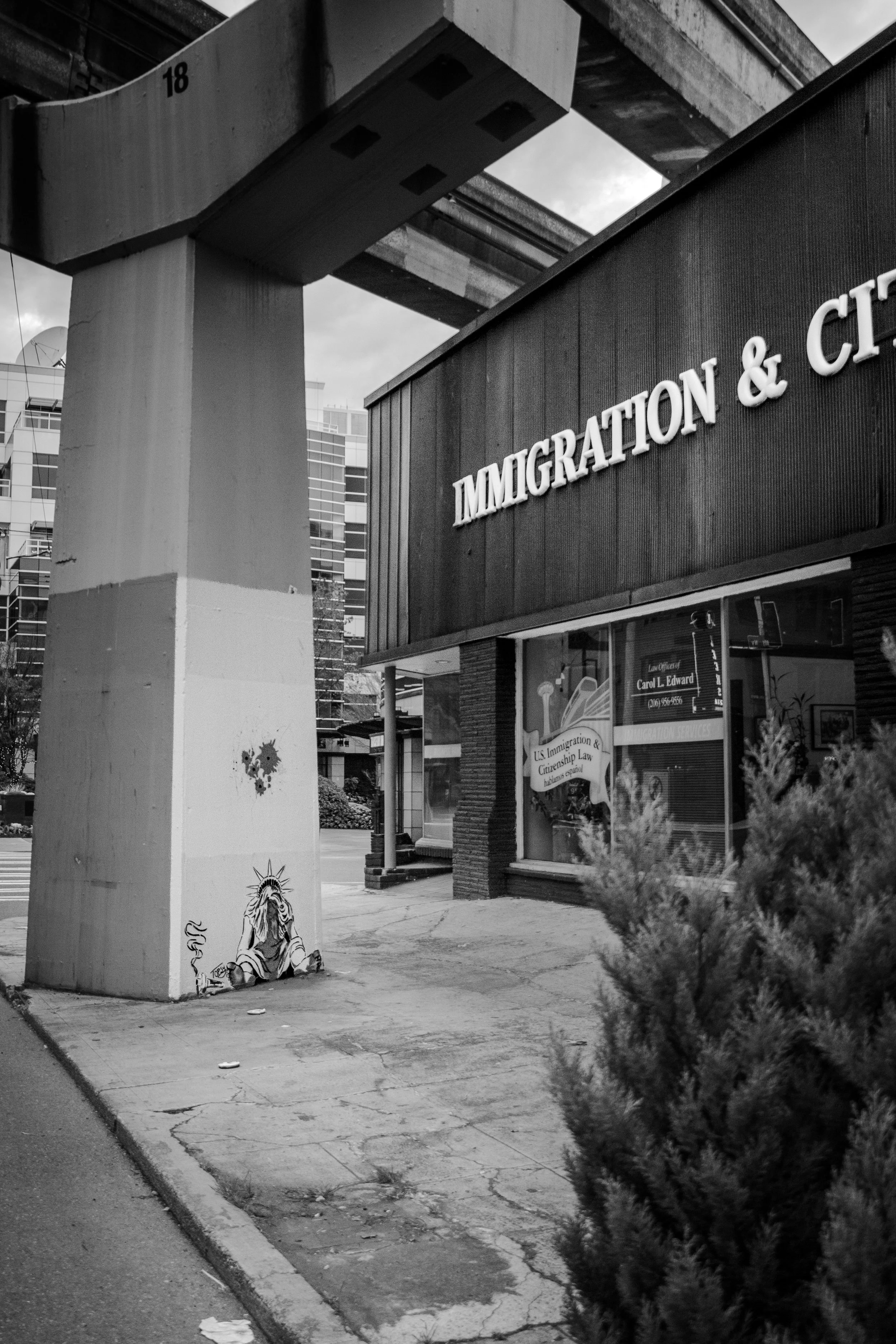 A black and white photo of a city street corner with a building sign reading 'IMMIGRATION & C' and graffiti artwork of a person with a crown and a lion on the pillar supporting an elevated train track overhead.