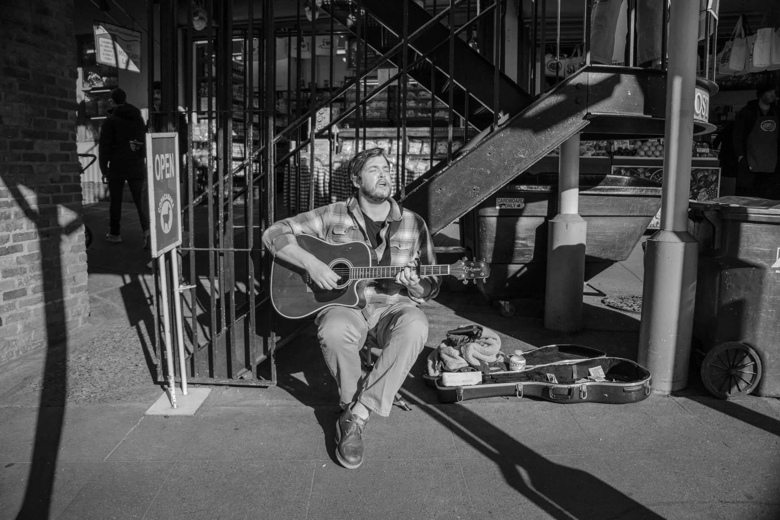 A street musician playing guitar and singing outdoors near a market entrance. The musician is sitting on a small stool with a guitar case open in front, possibly collecting tips. The scene is in black and white.