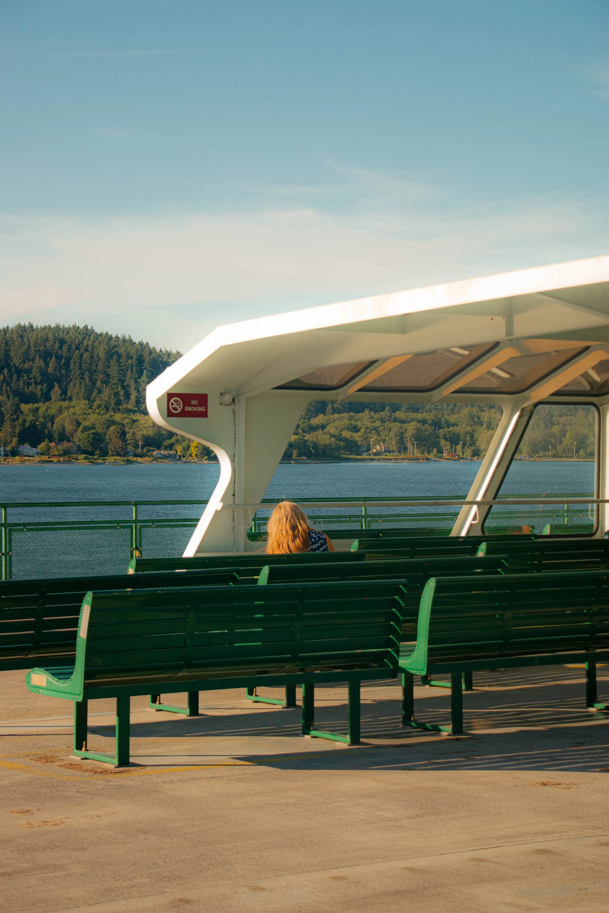 A woman with long red hair sitting alone on a dark green bench on a ferry overlooking a body of water and forested shoreline under a clear blue sky.