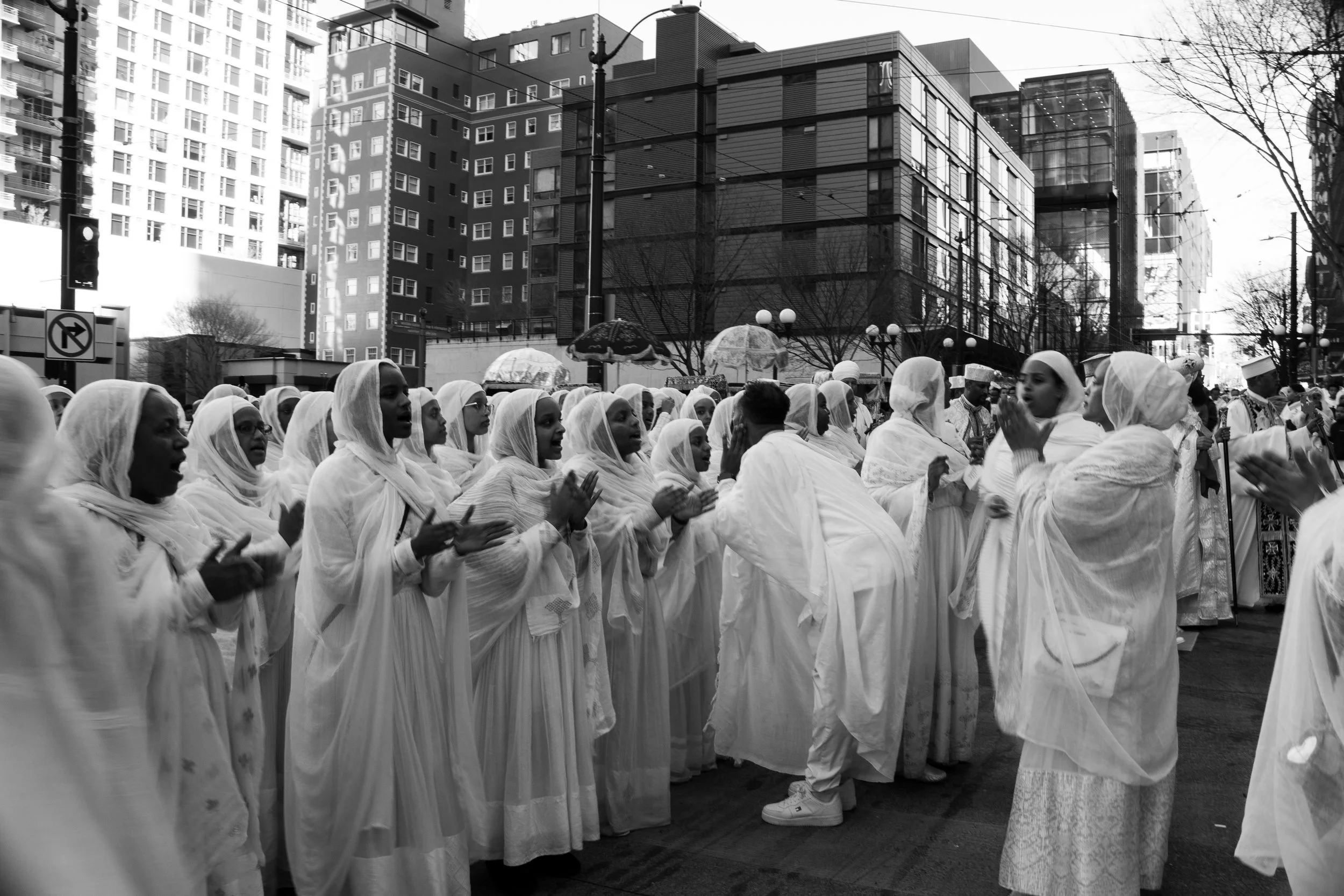 A group of people dressed in white robes, participating in a religious or cultural procession in an urban area, with modern buildings and trees in the background.