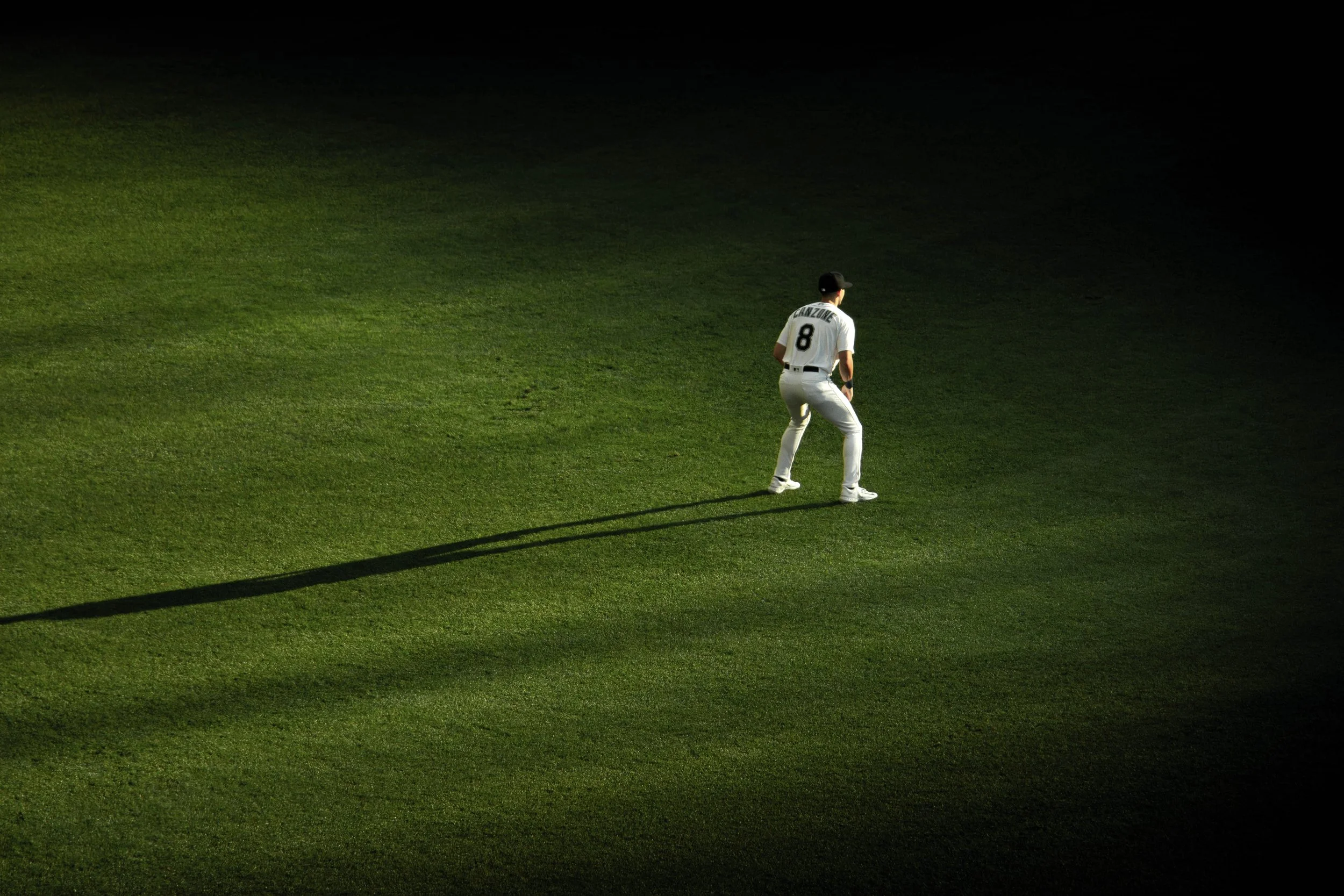 A baseball player standing on a grass field, shadow cast long on the ground, with a dark background.