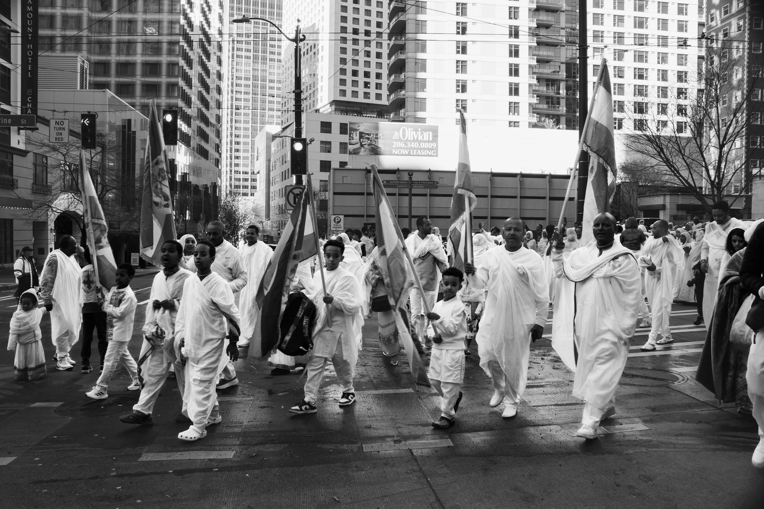 People in traditional white clothing participating in a parade on a city street, carrying flags, with tall buildings and traffic lights in the background.
