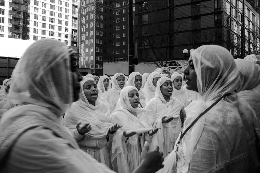 A group of women and girls dressed in white shawls and traditional attire, singing or chanting together on a city street with tall buildings in the background.