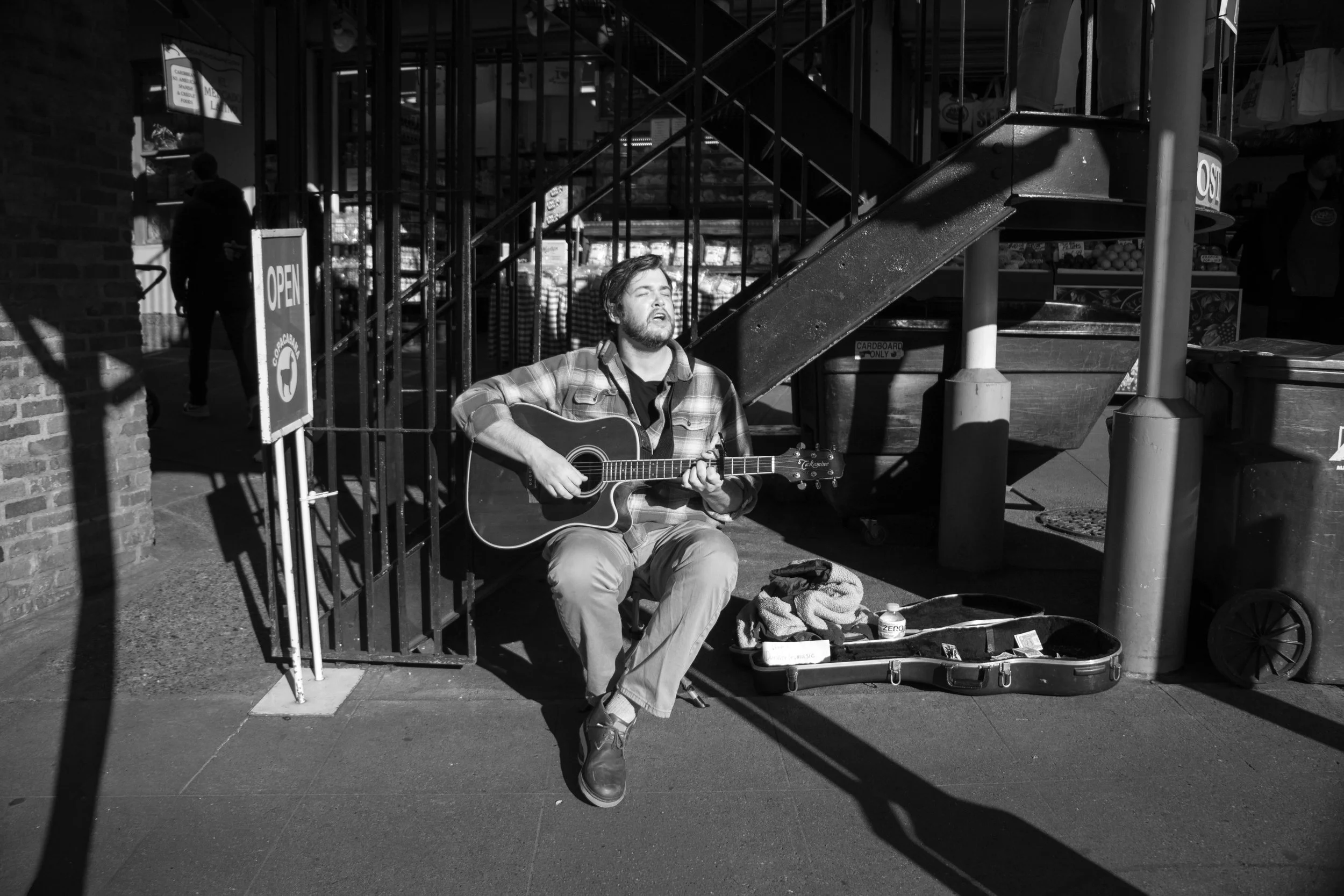 A man sits on the sidewalk playing an acoustic guitar with his eyes closed, surrounded by a small open guitar case with some belongings and money inside. The scene is outdoors near a store with metal stairs, trash bins, and another person walking in 