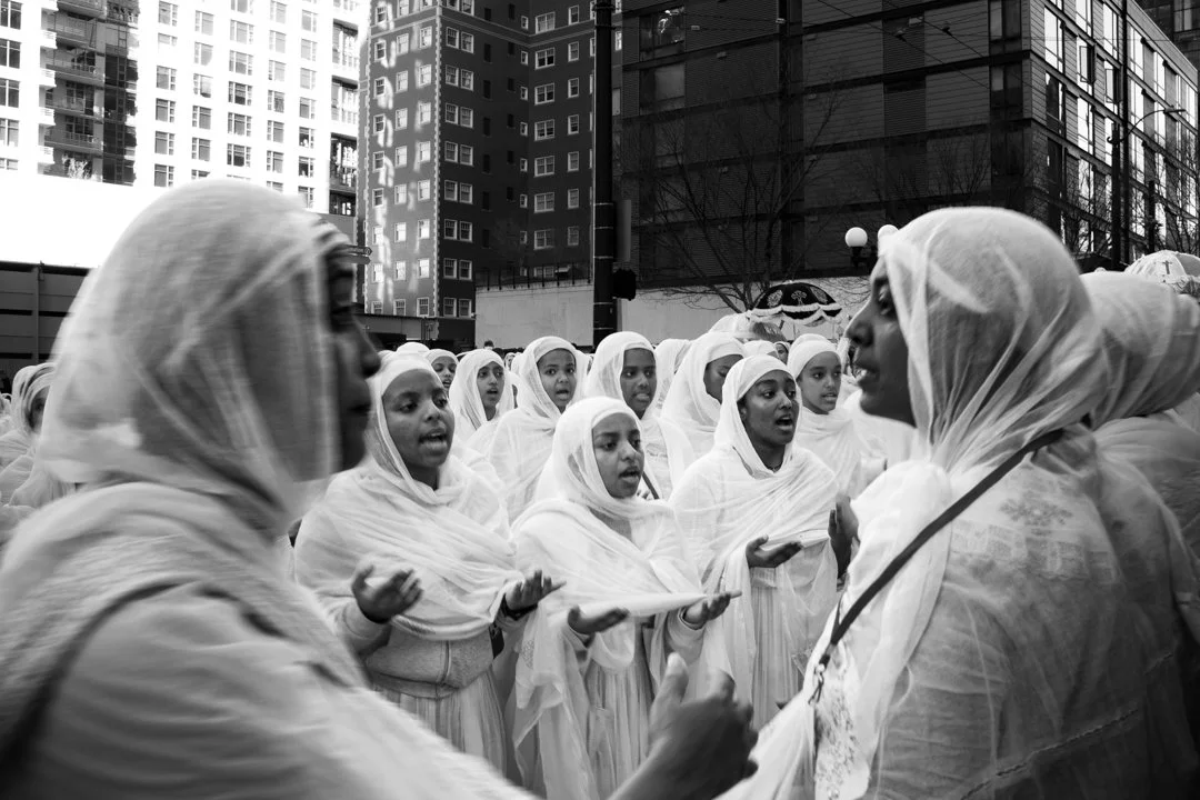 A group of women and girls dressed in white traditional clothing and headscarves gather and sing outdoors in an urban setting with tall buildings in the background.