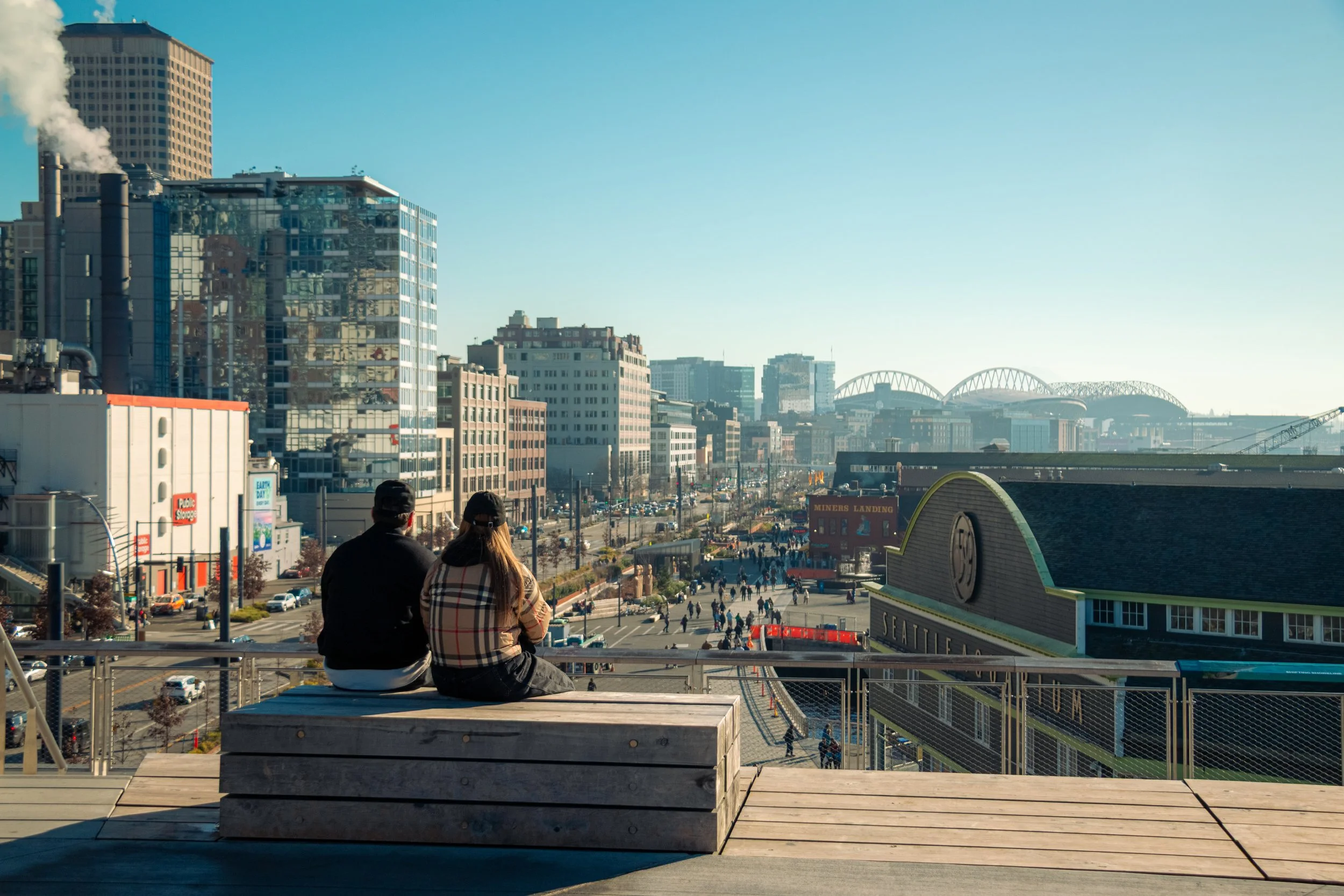 Two people sitting on a wooden bench overlooking downtown Seattle with a view of the city skyline and the Mariners baseball stadium.