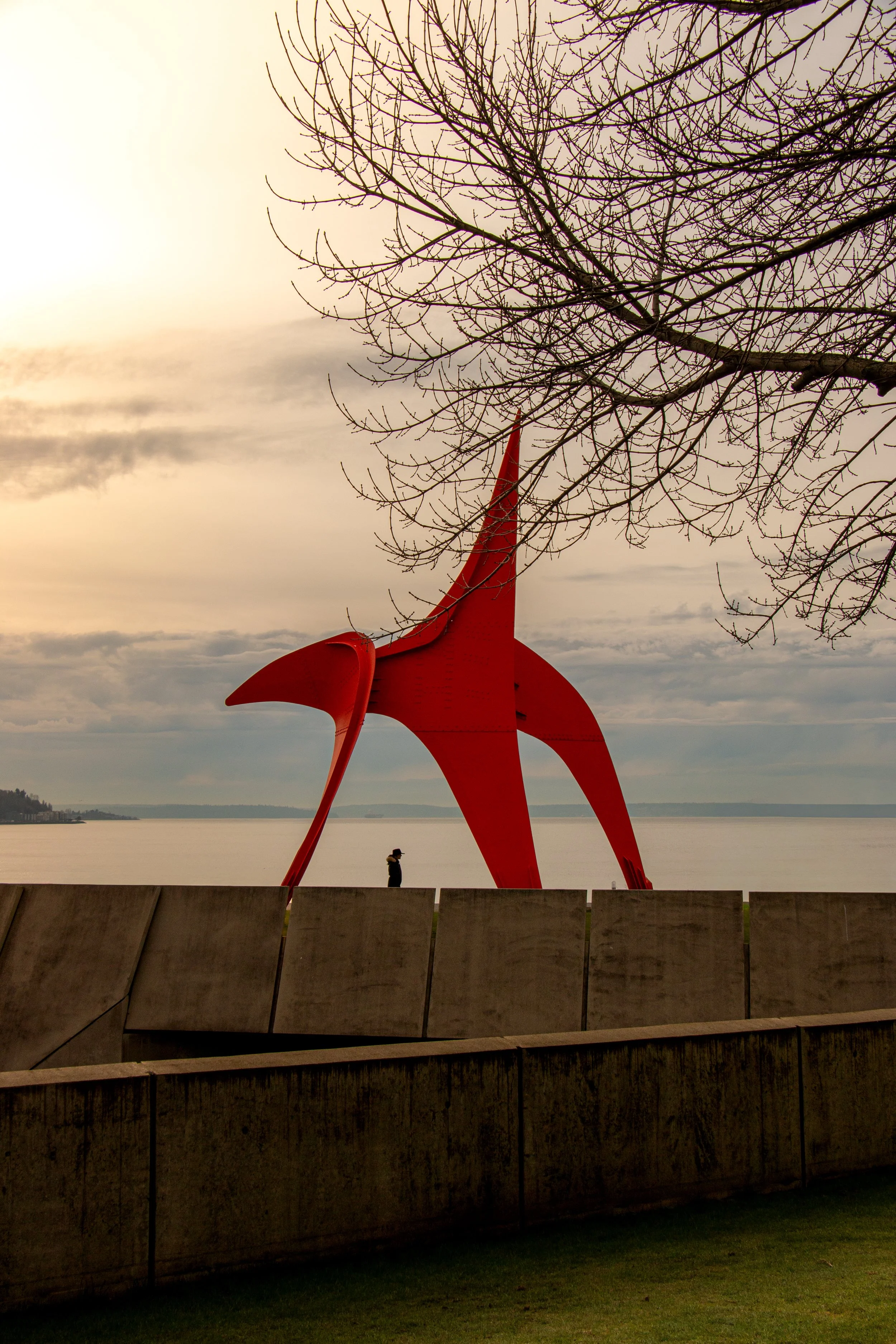 A person walking along a waterfront promenade with a large red abstract sculpture and a leafless tree in the foreground, overlooking calm water and cloudy sky.