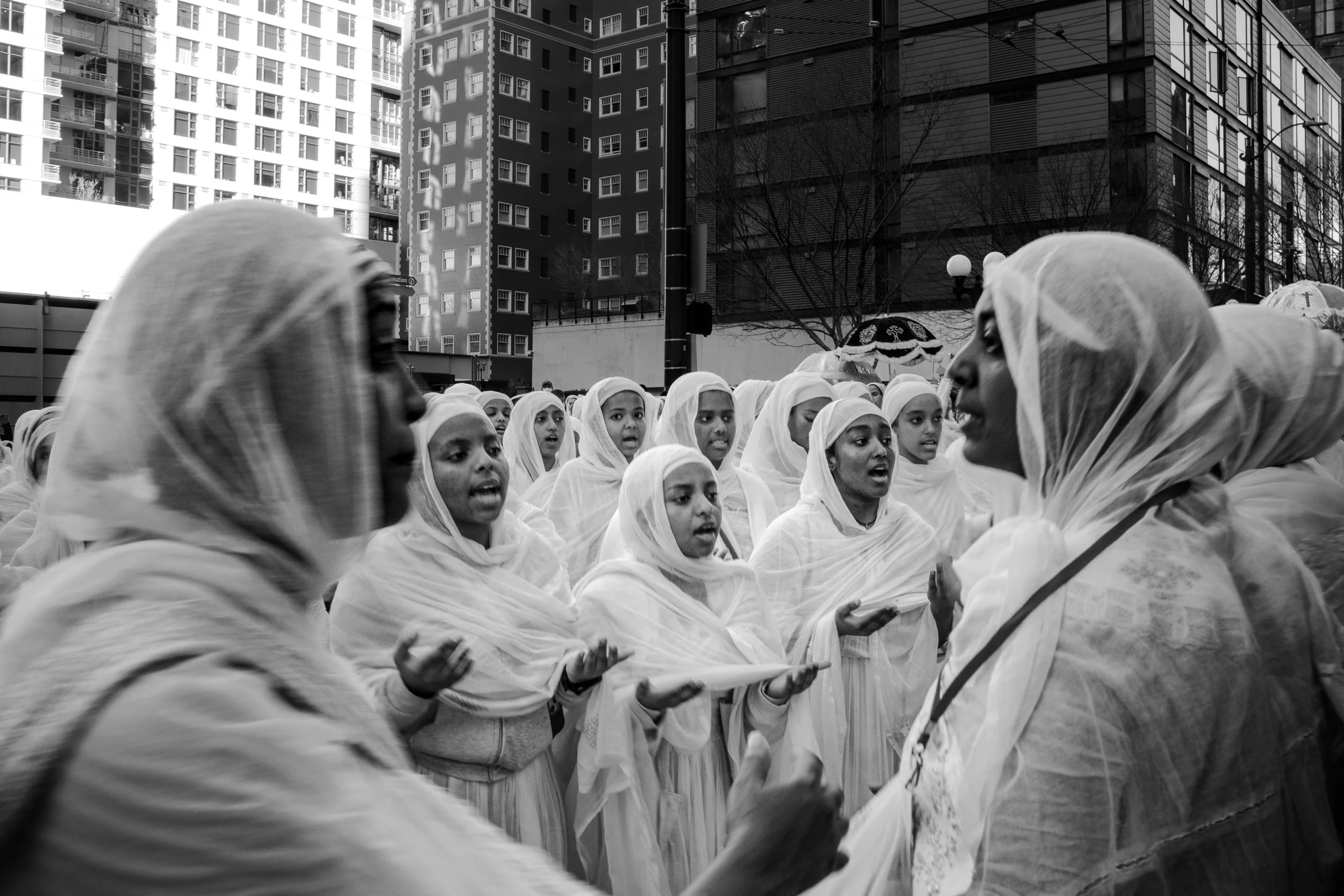 A group of women and girls dressed in white, standing close together outdoors in a city square, seemingly singing or speaking to a woman at the front, with tall buildings in the background.
